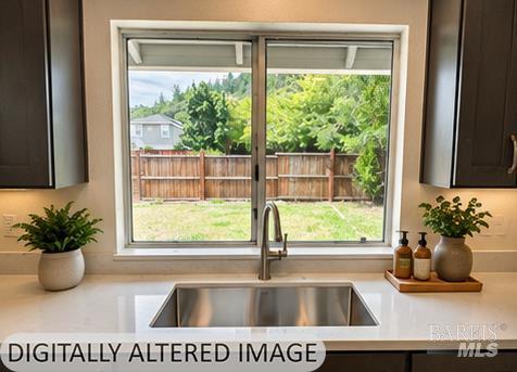 Kitchen sink beneath a wide window overlooking a fenced backyard, with dark cabinets and countertop plants.