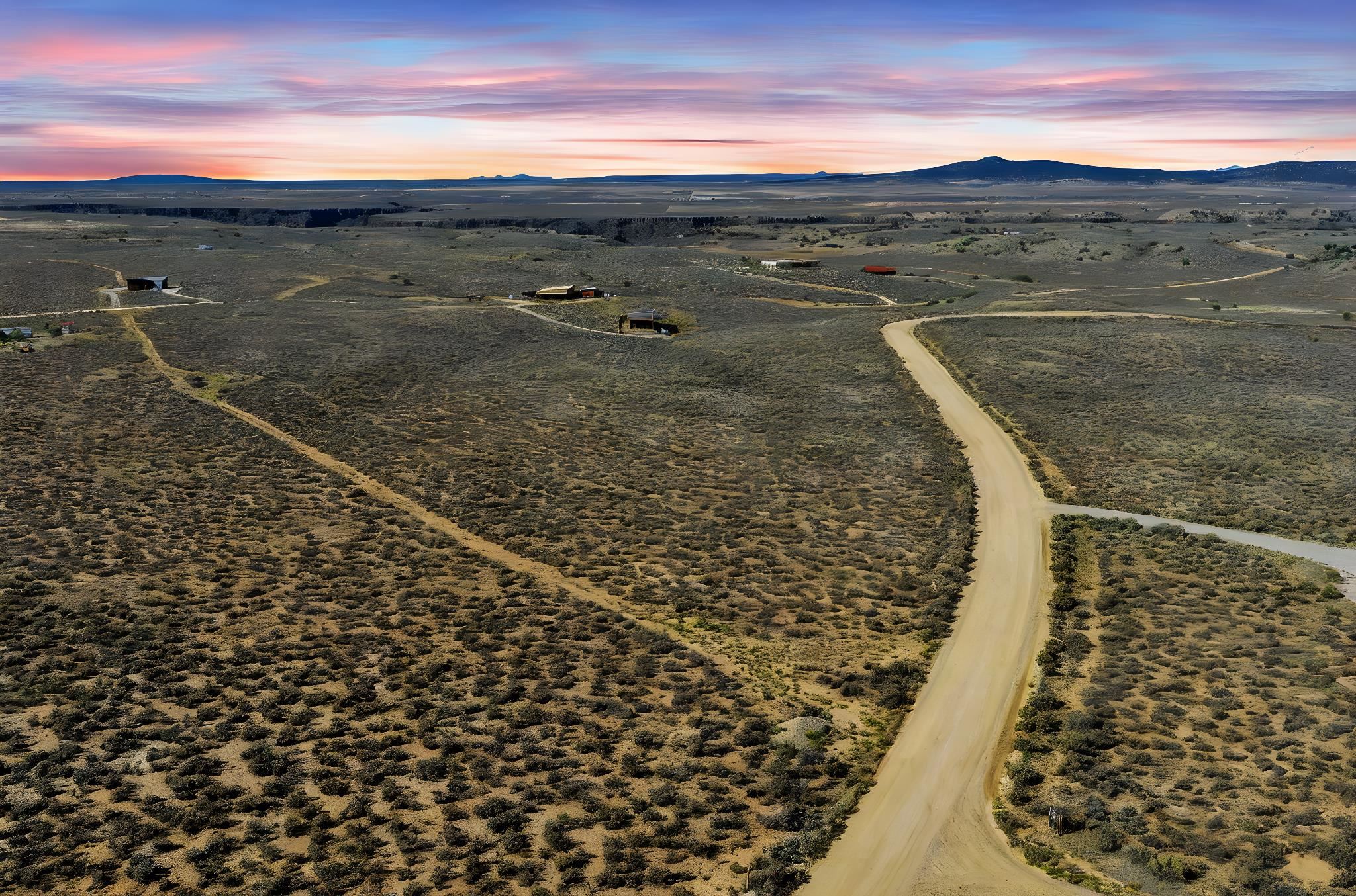 Aerial view of scrubland acreage with a winding dirt road and distant mountains at dusk.