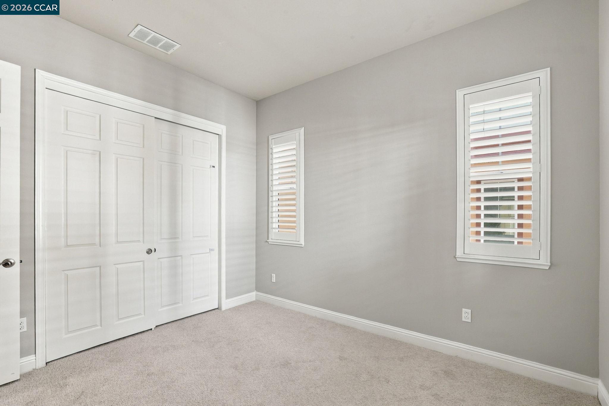 Empty bedroom with beige carpet, white closet doors, and two shuttered windows.