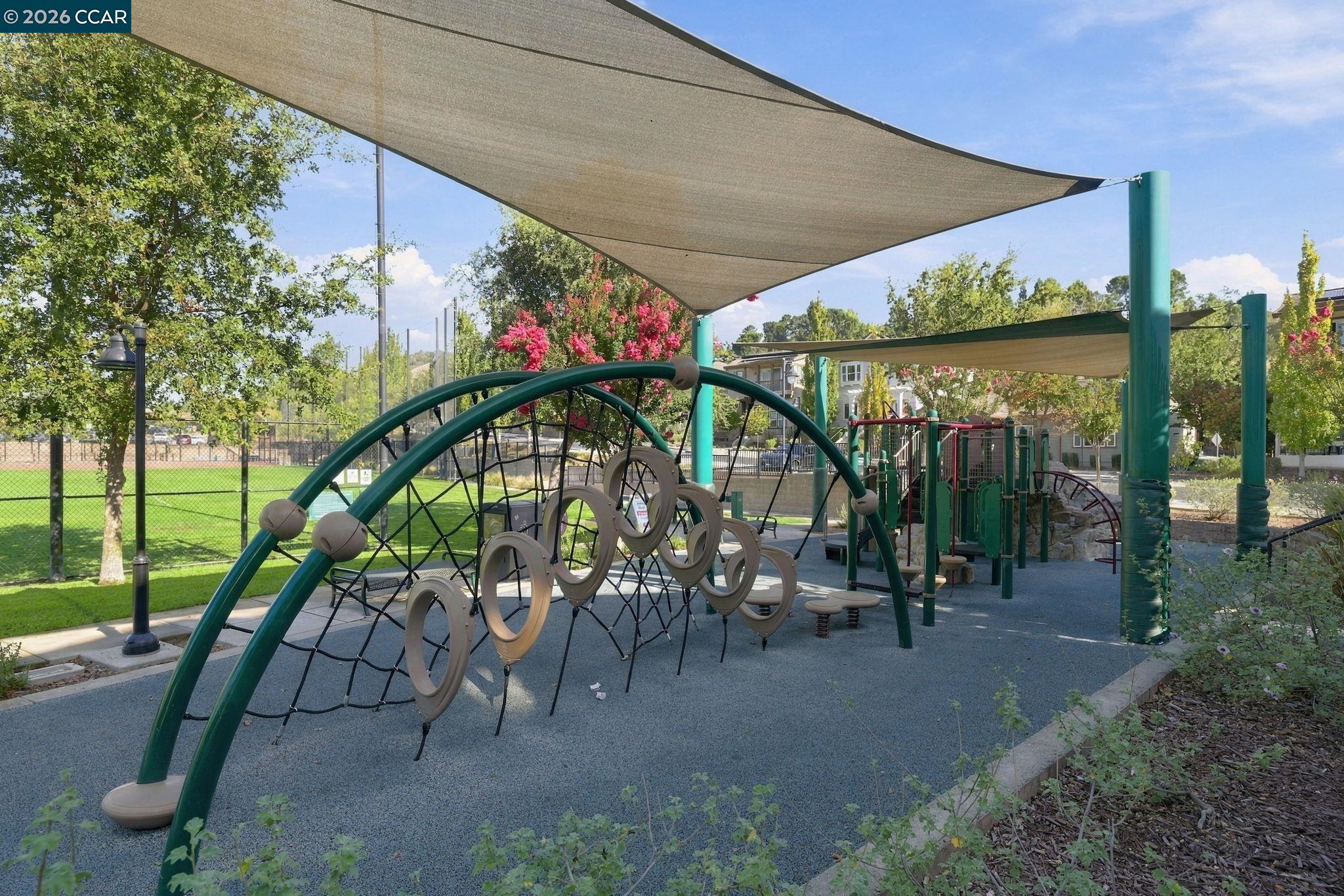 Outdoor playground with climbing equipment beneath shade sails, surrounded by trees and a fenced lawn.