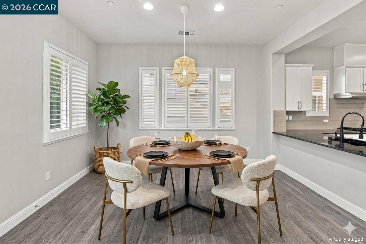 Dining area with a round table set for four, woven pendant light, shuttered windows, and adjoining white kitchen.