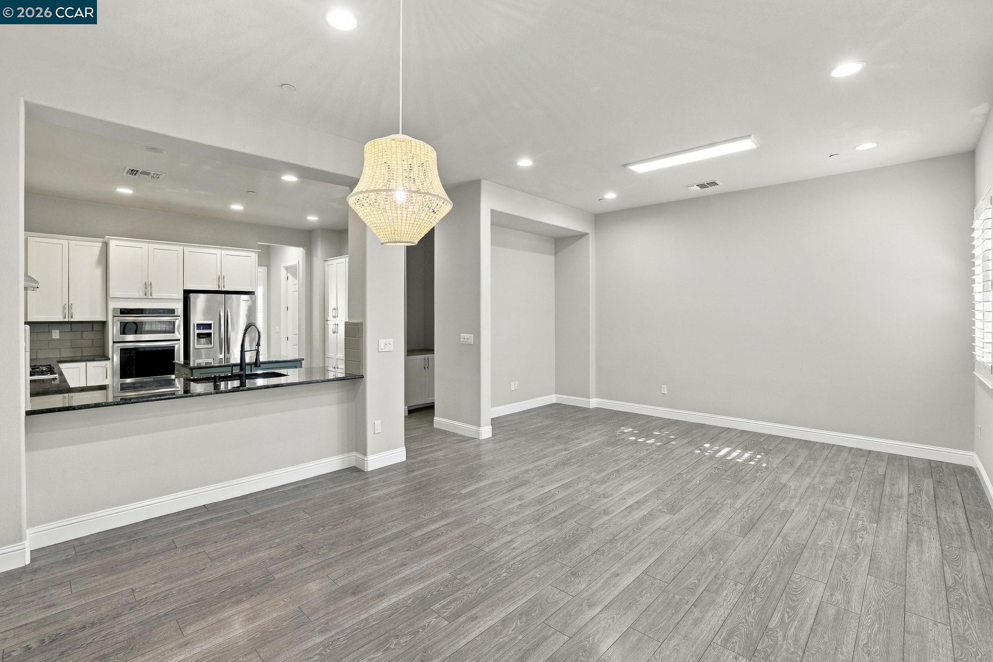 Empty dining area with gray plank flooring, a woven pendant light, and an adjoining white kitchen.