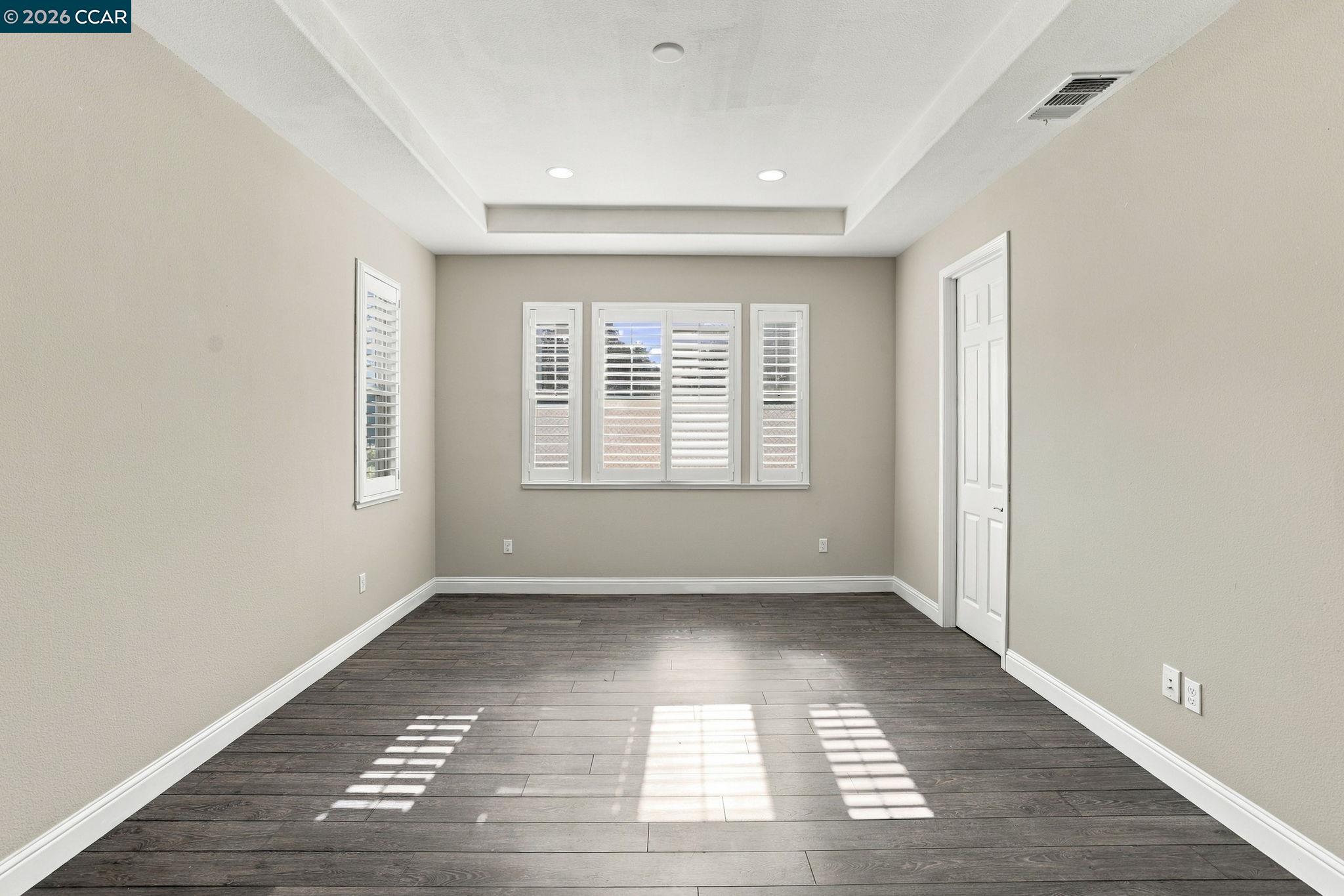 Empty room with dark plank flooring, plantation-shuttered windows, and a recessed tray ceiling.