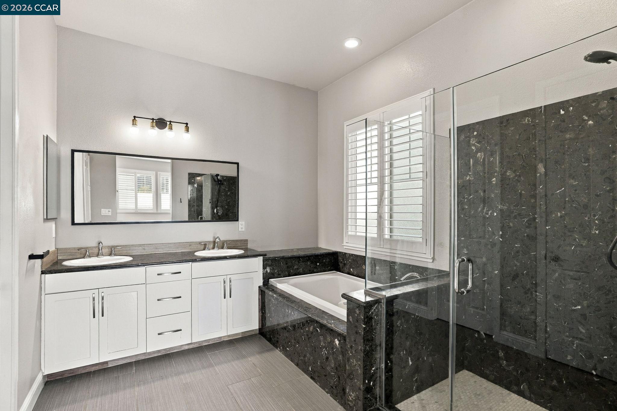 Bathroom with double vanity, dark stone bathtub surround, and glass-enclosed shower.