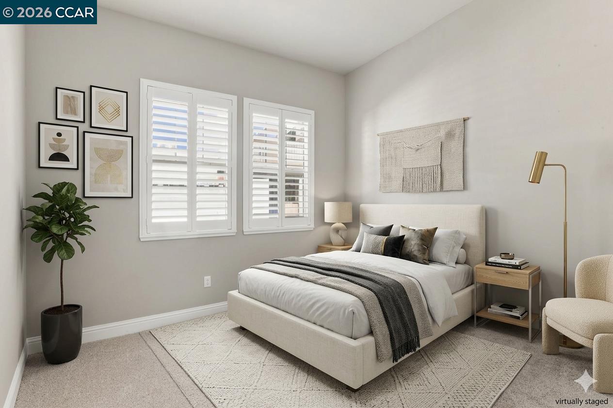 Staged bedroom with a cream upholstered bed, white plantation shutters, framed art, and a potted plant.