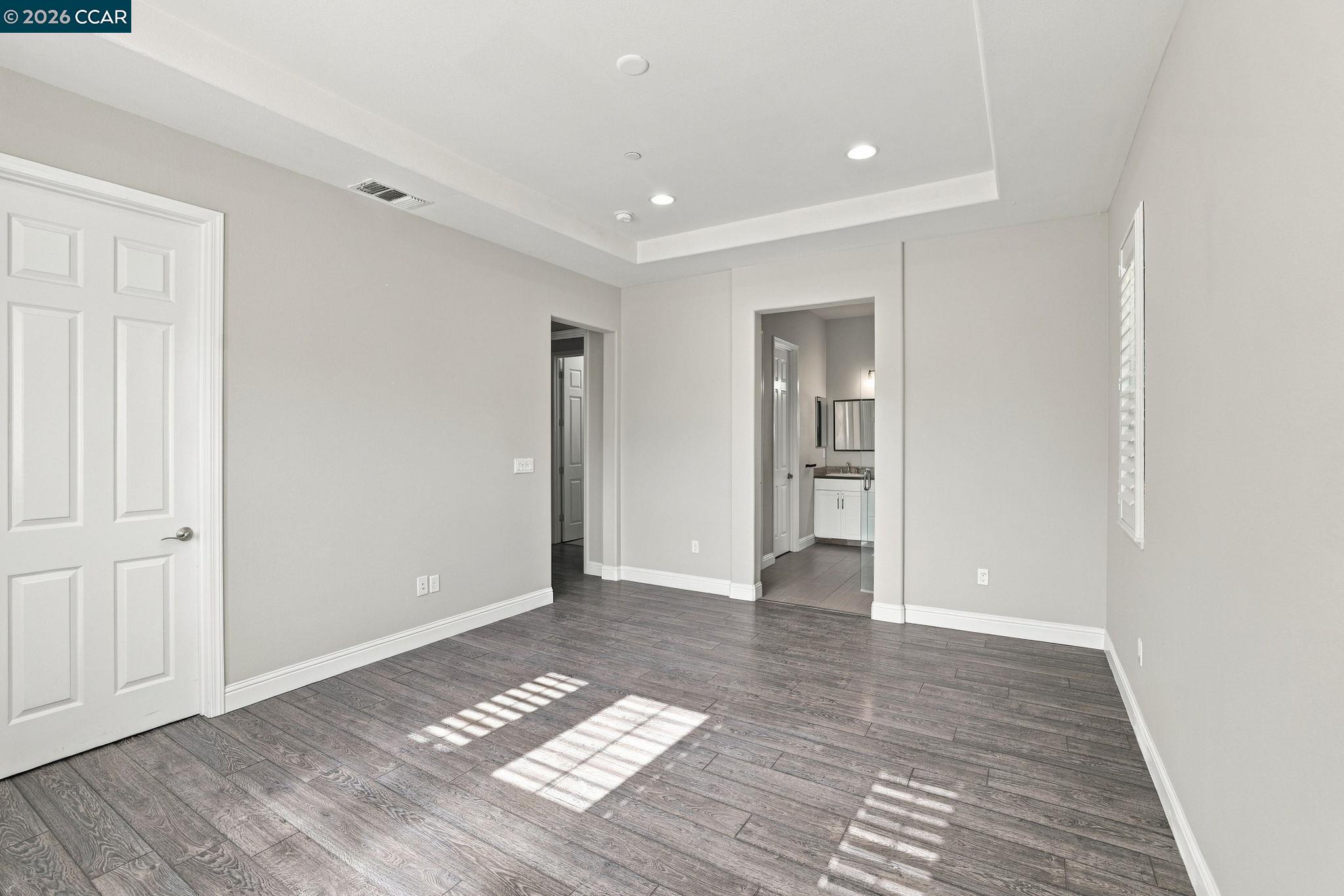 Empty bedroom with gray plank flooring and a tray ceiling, opening to a bathroom with a glass shower.