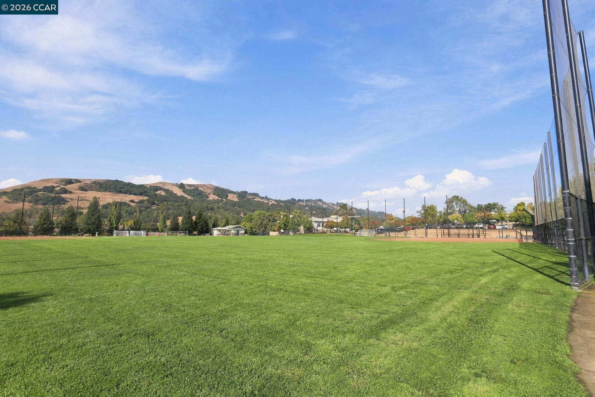 Wide green sports field with baseball diamonds, protective fencing, and rolling hills in the background.