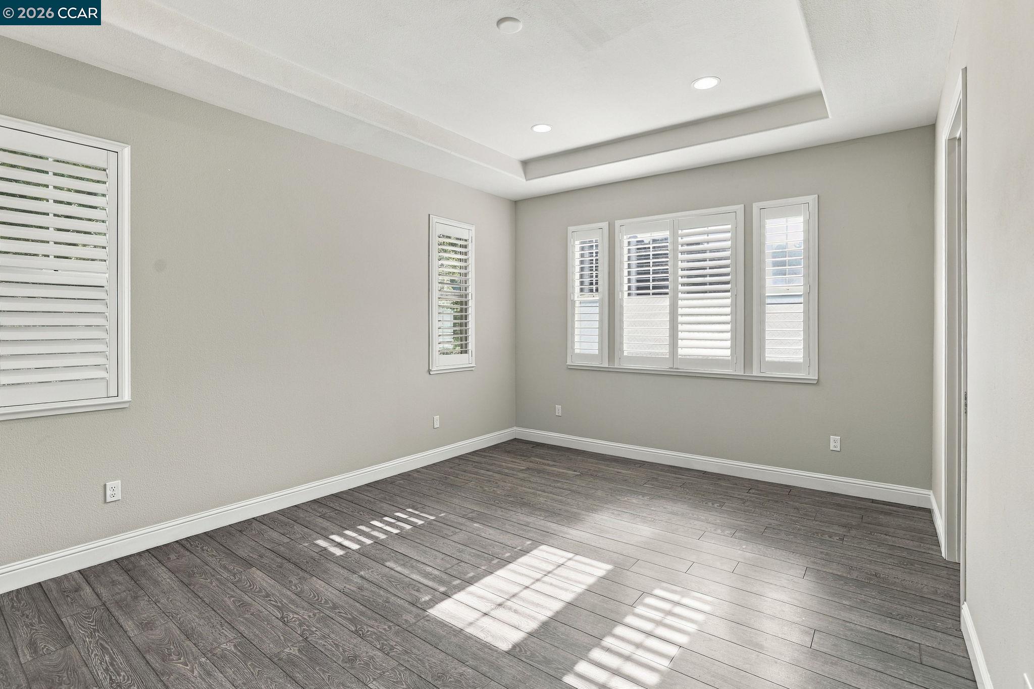 Empty room with gray plank floors, a tray ceiling, and shuttered windows.
