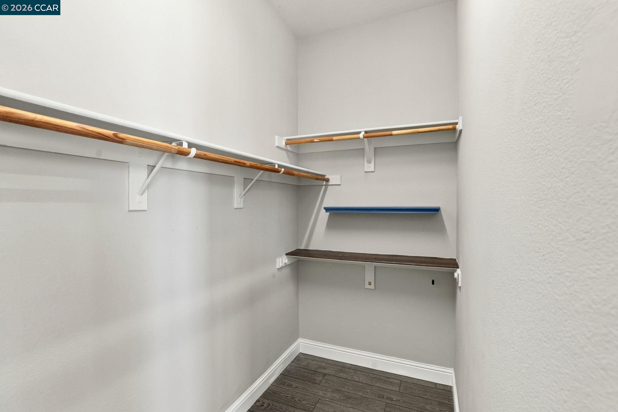 Walk-in closet with wooden hanging rods, wall shelves, and dark wood-look flooring.