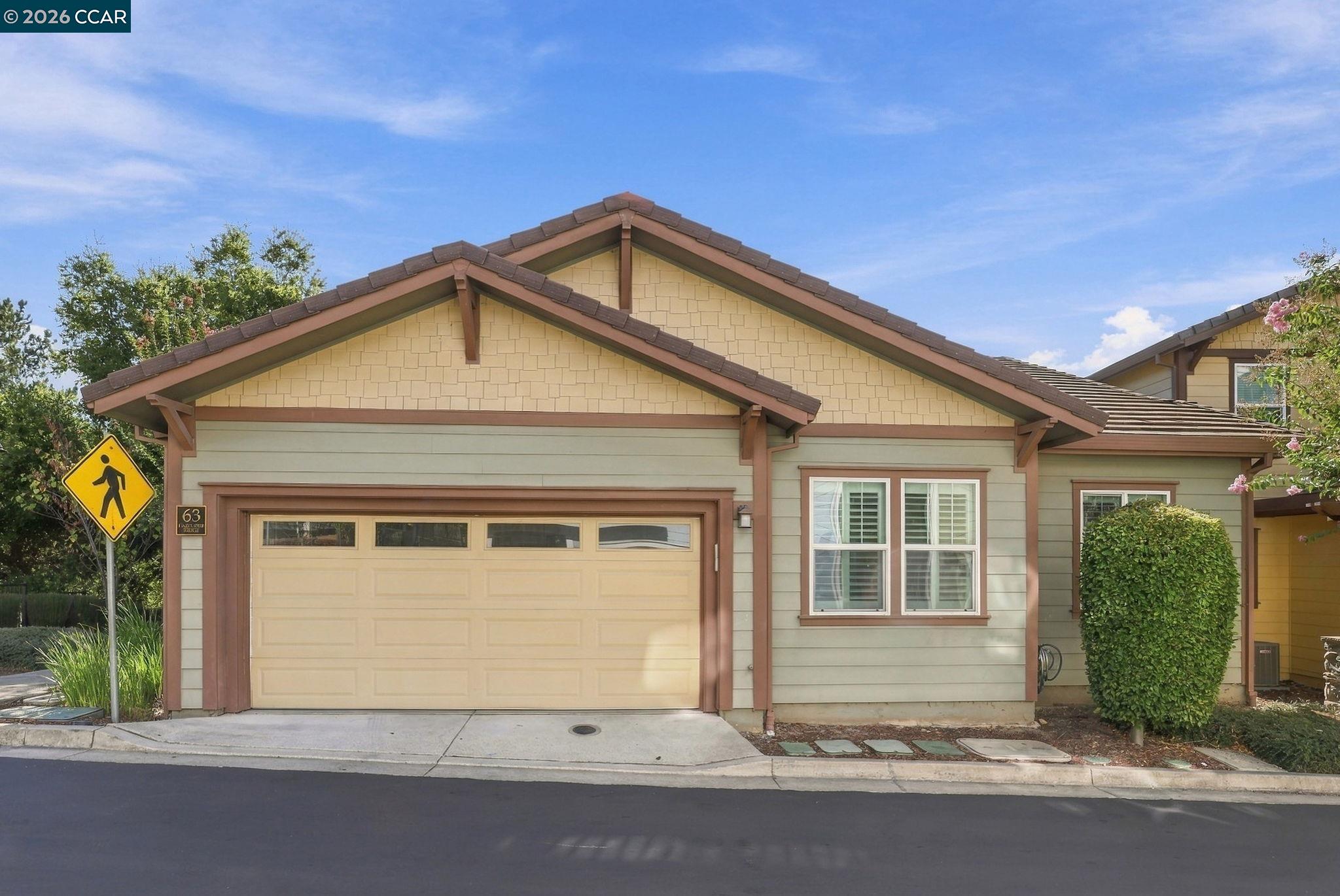 Single-story home with a wide double garage door, sage-green siding, and a pedestrian crossing sign.