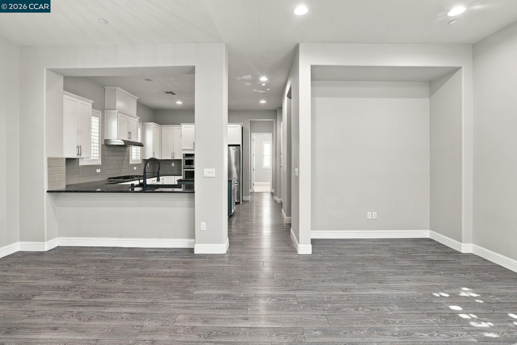 Unfurnished living area with gray plank flooring, open to a white-cabinet kitchen.