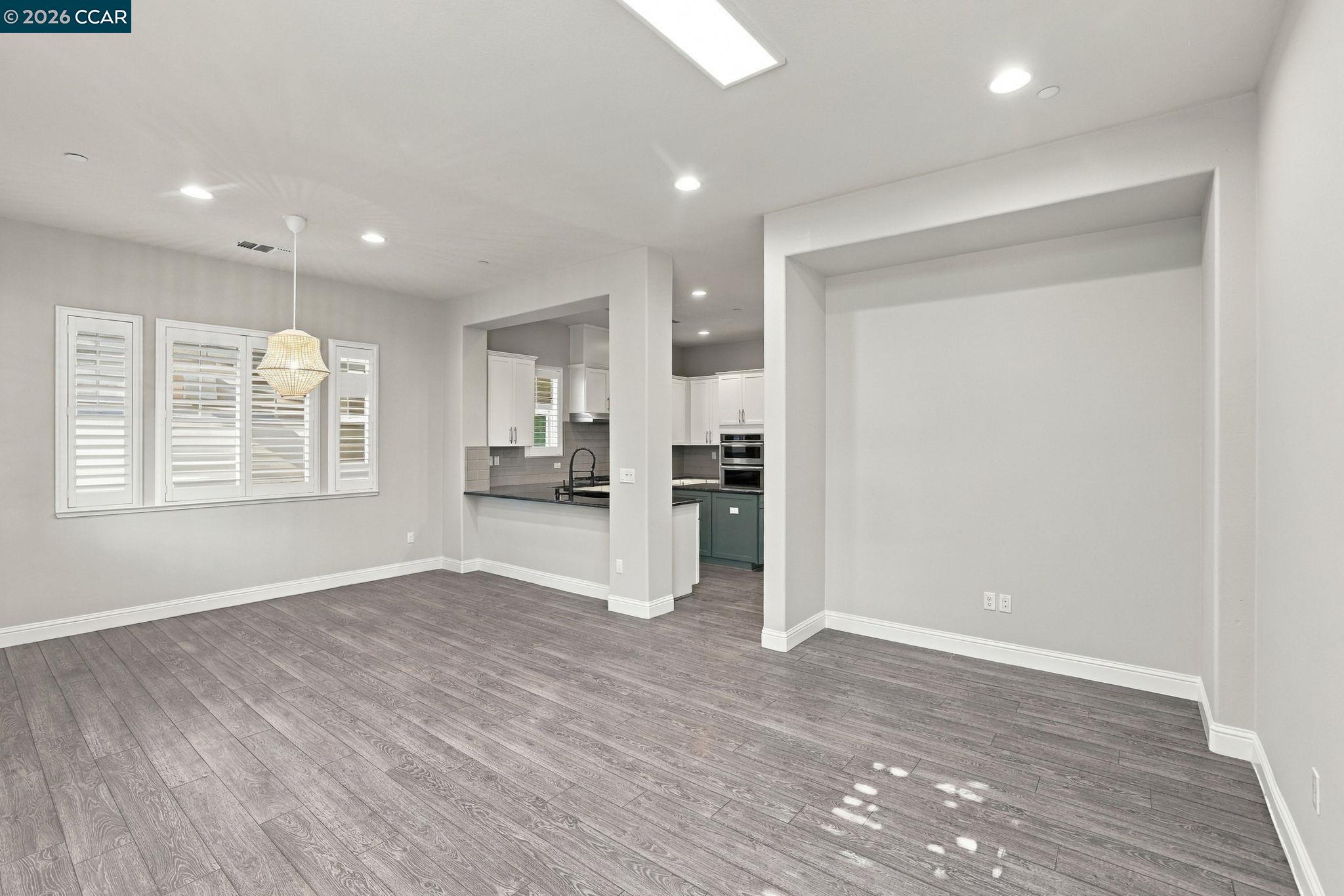 Empty dining area with gray plank flooring, plantation shutters, and an open view into the kitchen.