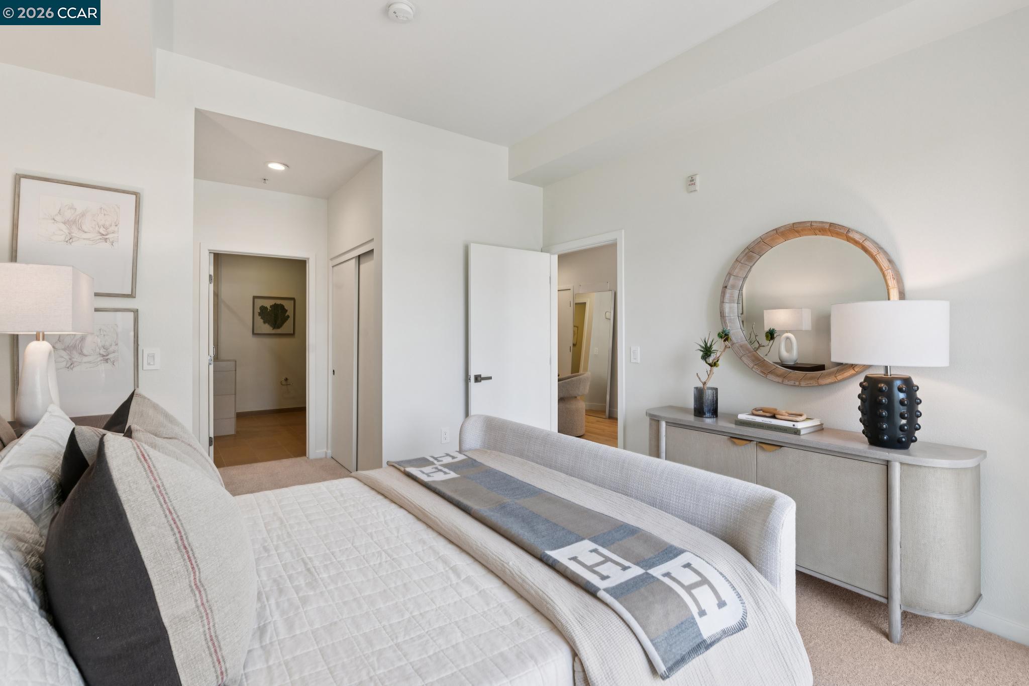 Staged bedroom with layered gray and white bedding and a large round mirror.