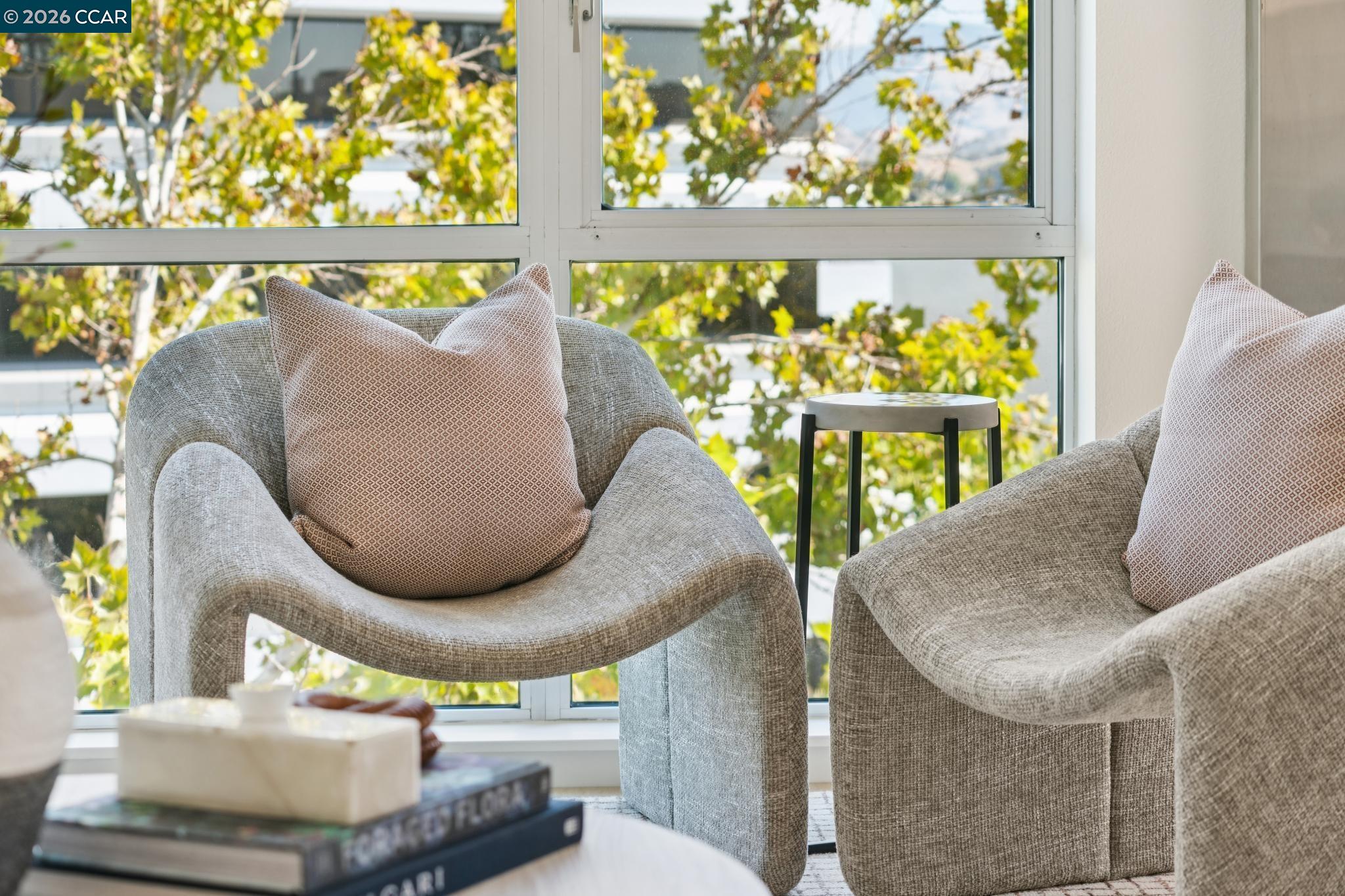 Living room with gray lounge chairs and patterned pillows beside a large window overlooking trees.