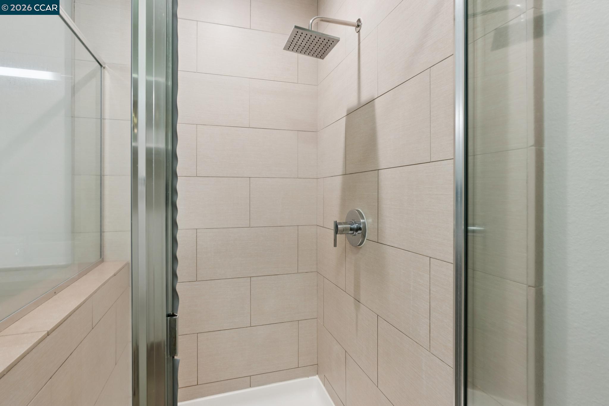 Walk-in shower with beige tile, a square rainfall showerhead, and glass doors.