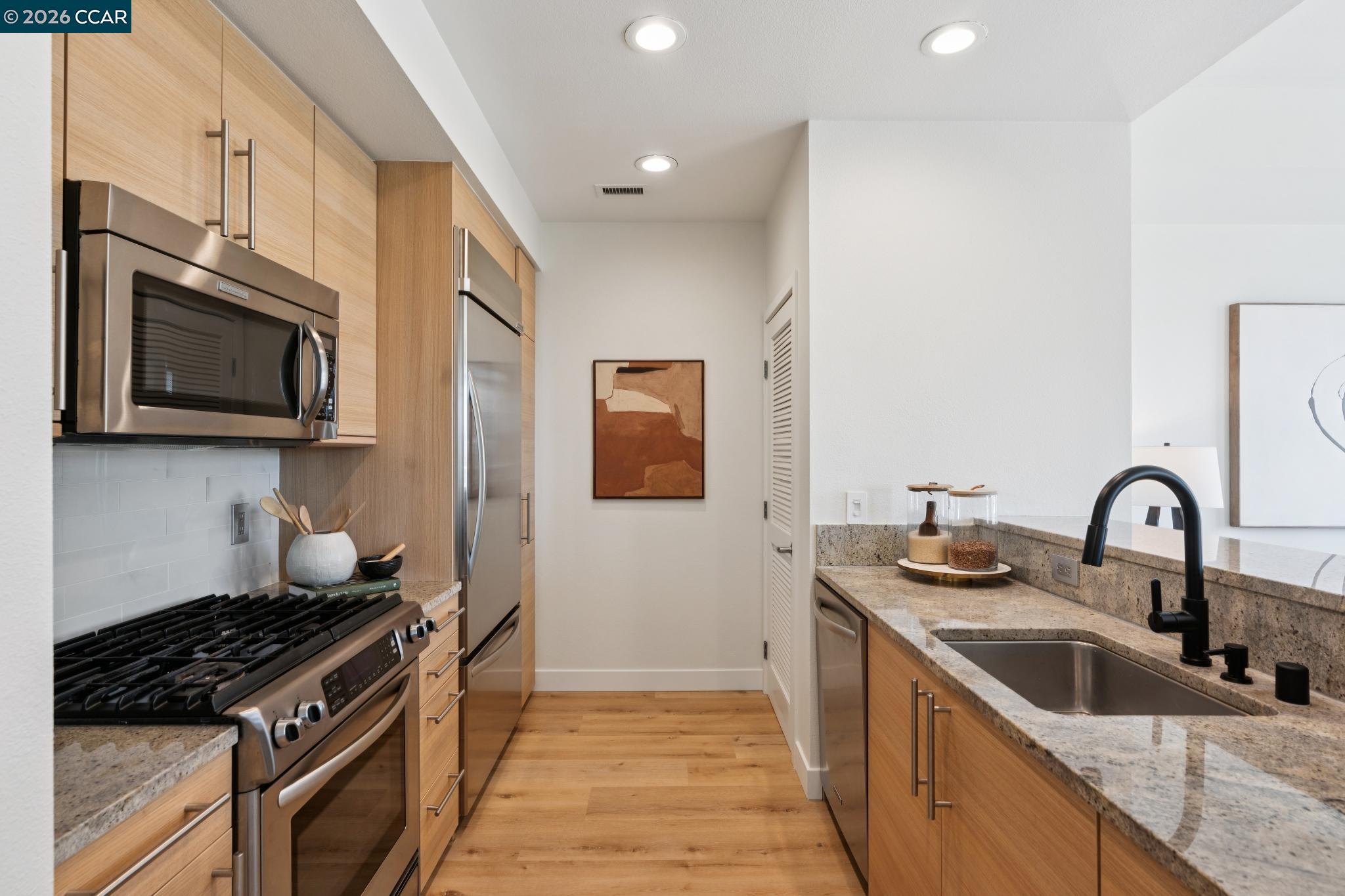 Modern galley kitchen with light wood cabinets, stainless steel appliances, and granite counters.