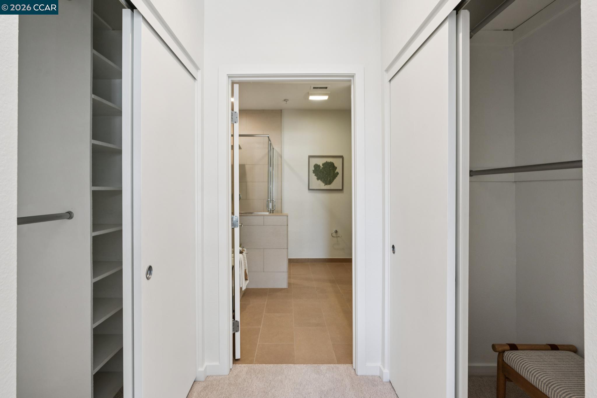 Walk-in closet with built-in shelves and hanging rails, opening to a tiled bathroom with a glass shower.