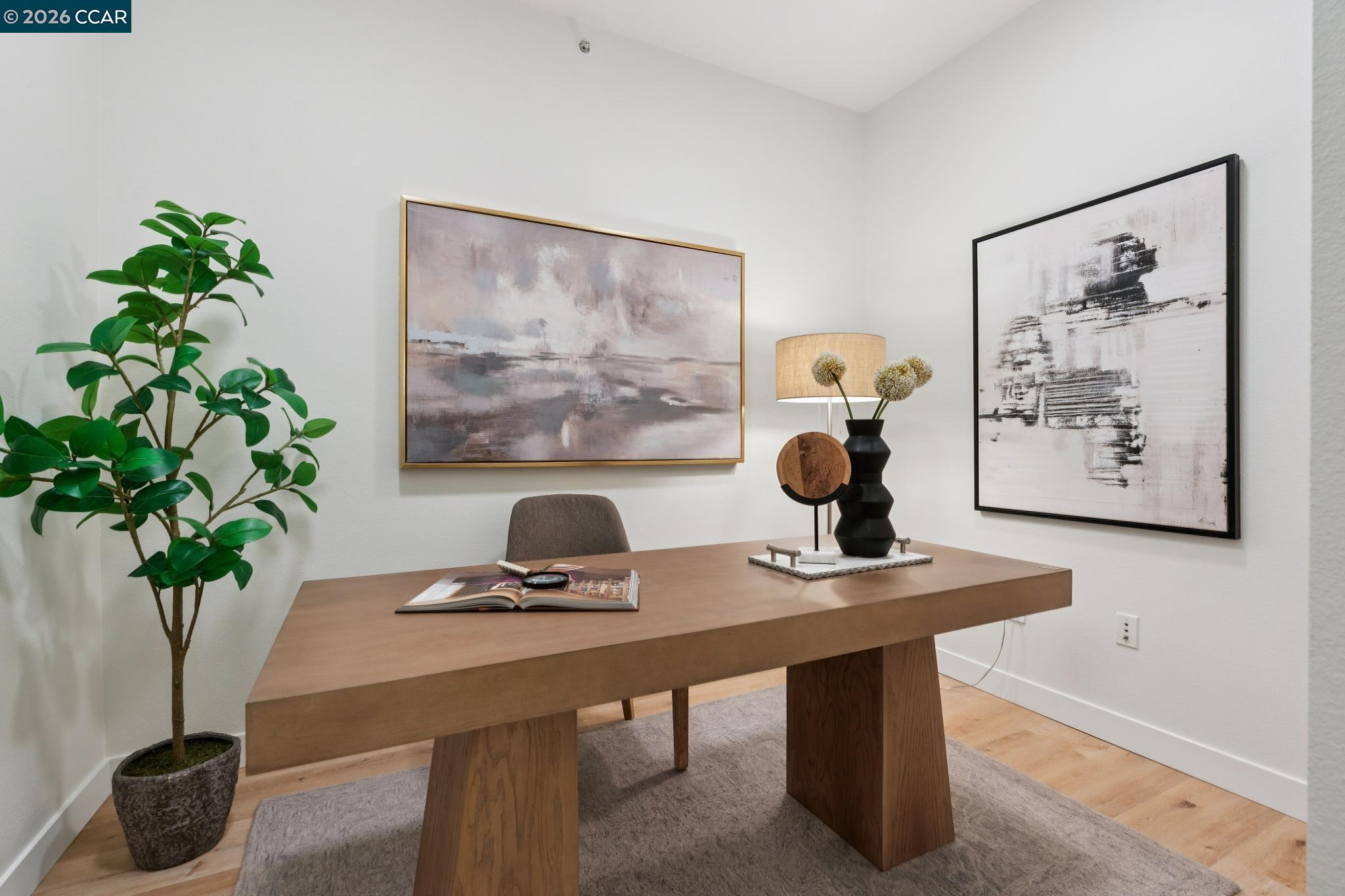 Staged home office with a broad wooden desk, potted tree, and two abstract paintings.