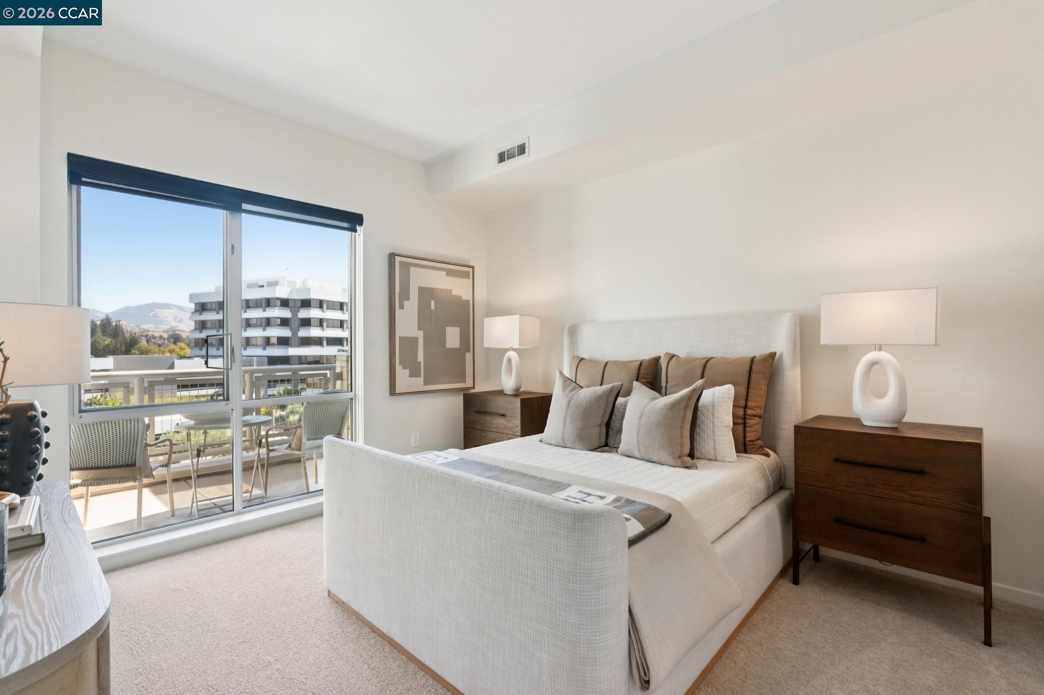 Staged bedroom with a king-size bed and balcony doors overlooking buildings and distant hills.