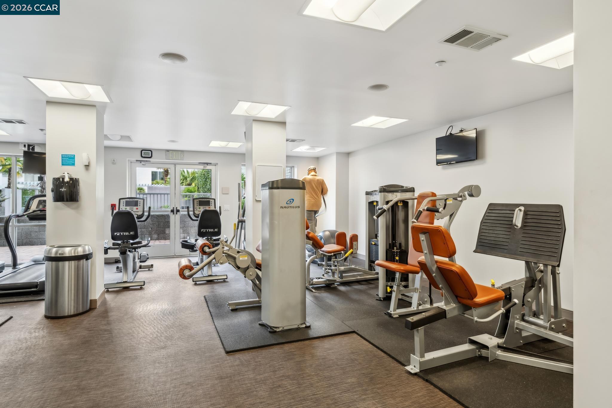 Fitness center with strength machines, recumbent bikes, and glass doors opening to greenery.