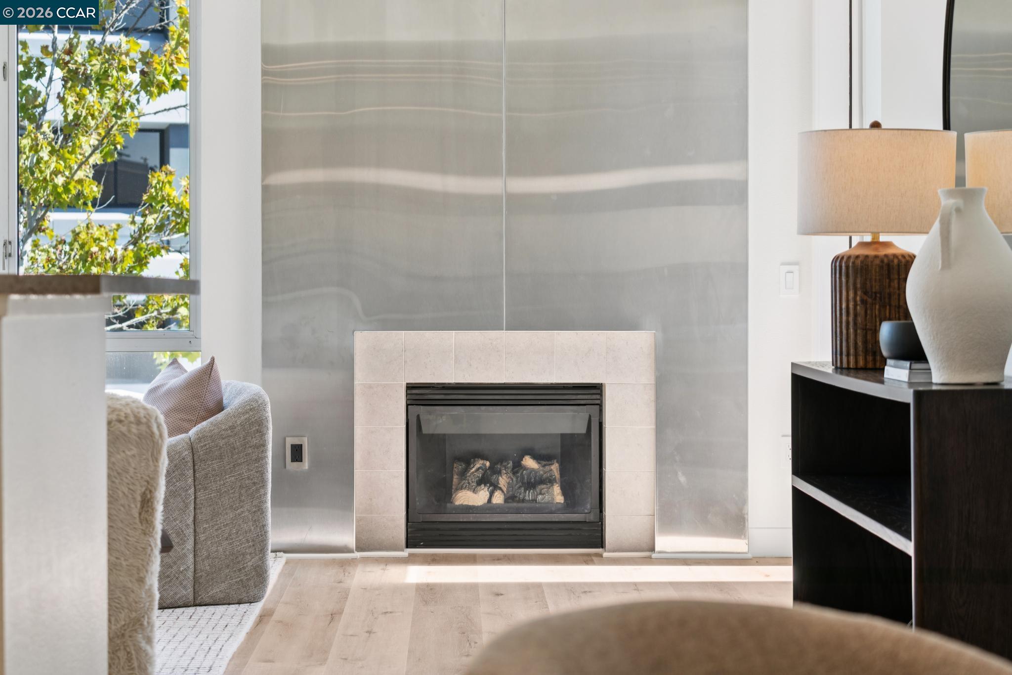 Living room with a modern fireplace, gray stone wall, pale wood floors, and a window overlooking leafy trees.