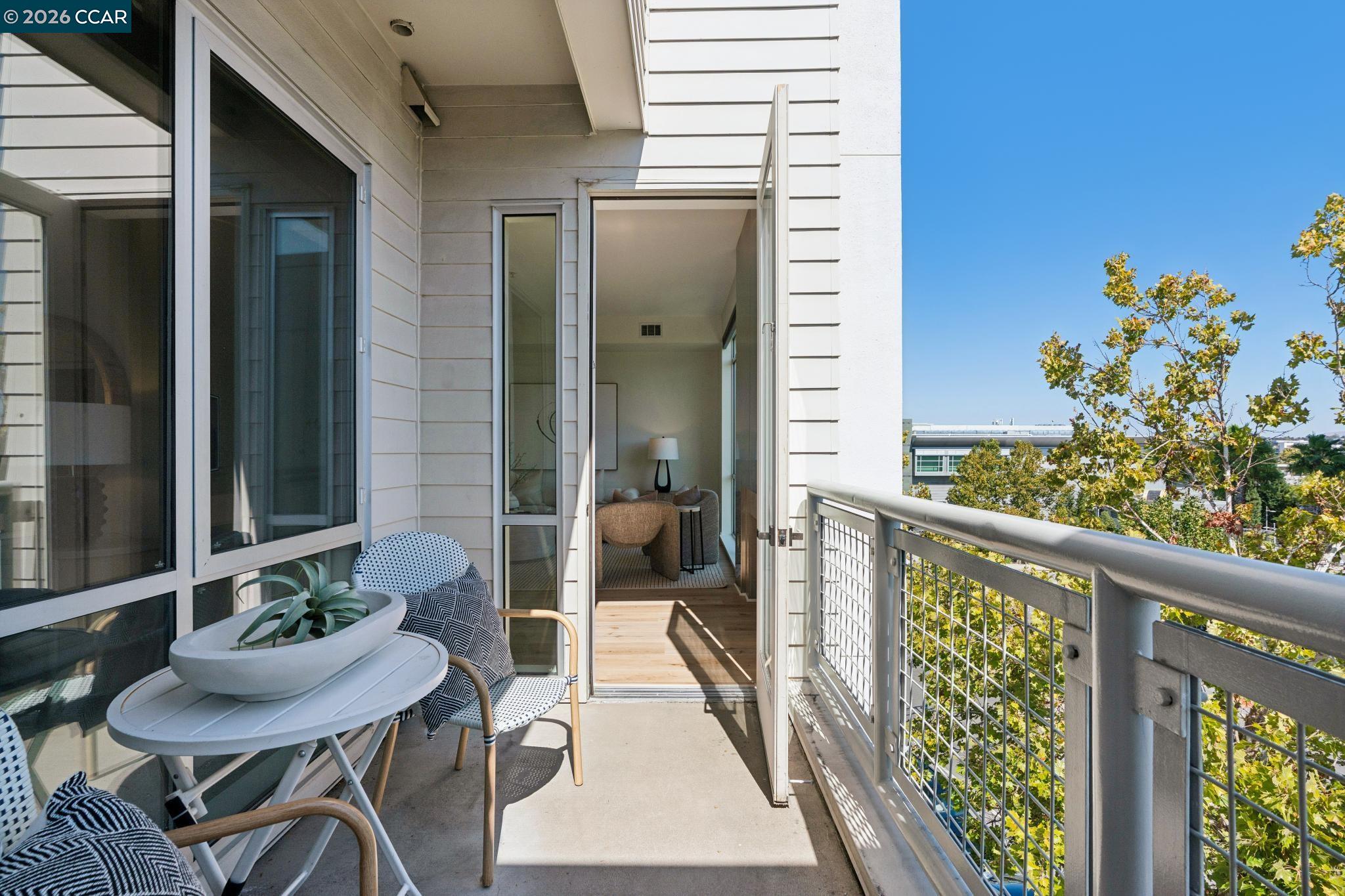 Furnished apartment balcony with seating, open glass door, and views of trees and nearby buildings.