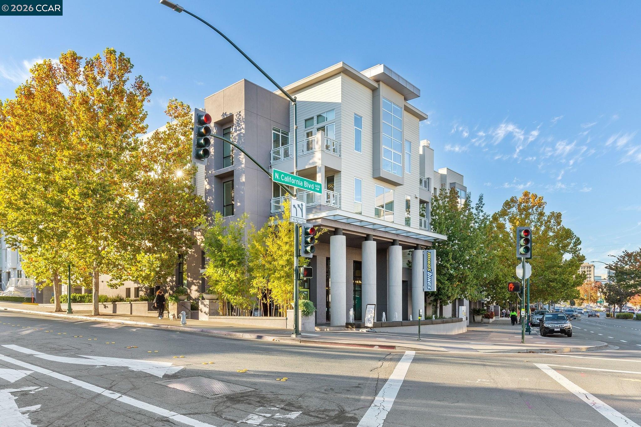 Modern multi-story condominium building with balconies and columns at a tree-lined street corner.