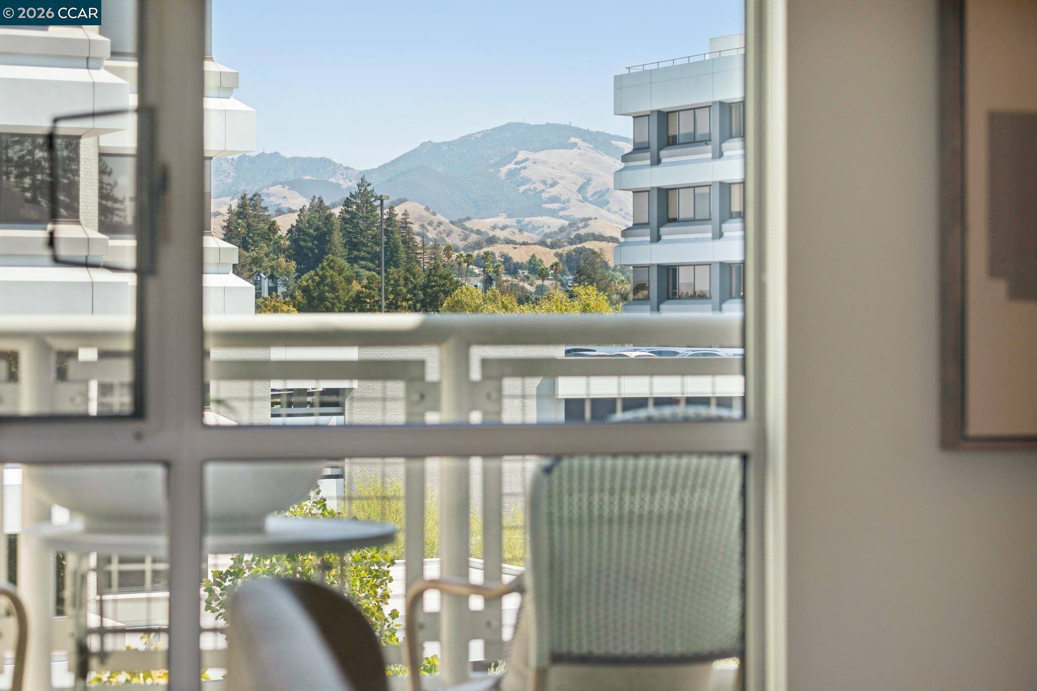 Balcony view of neighboring buildings, leafy trees, and rolling hills beyond the railing.