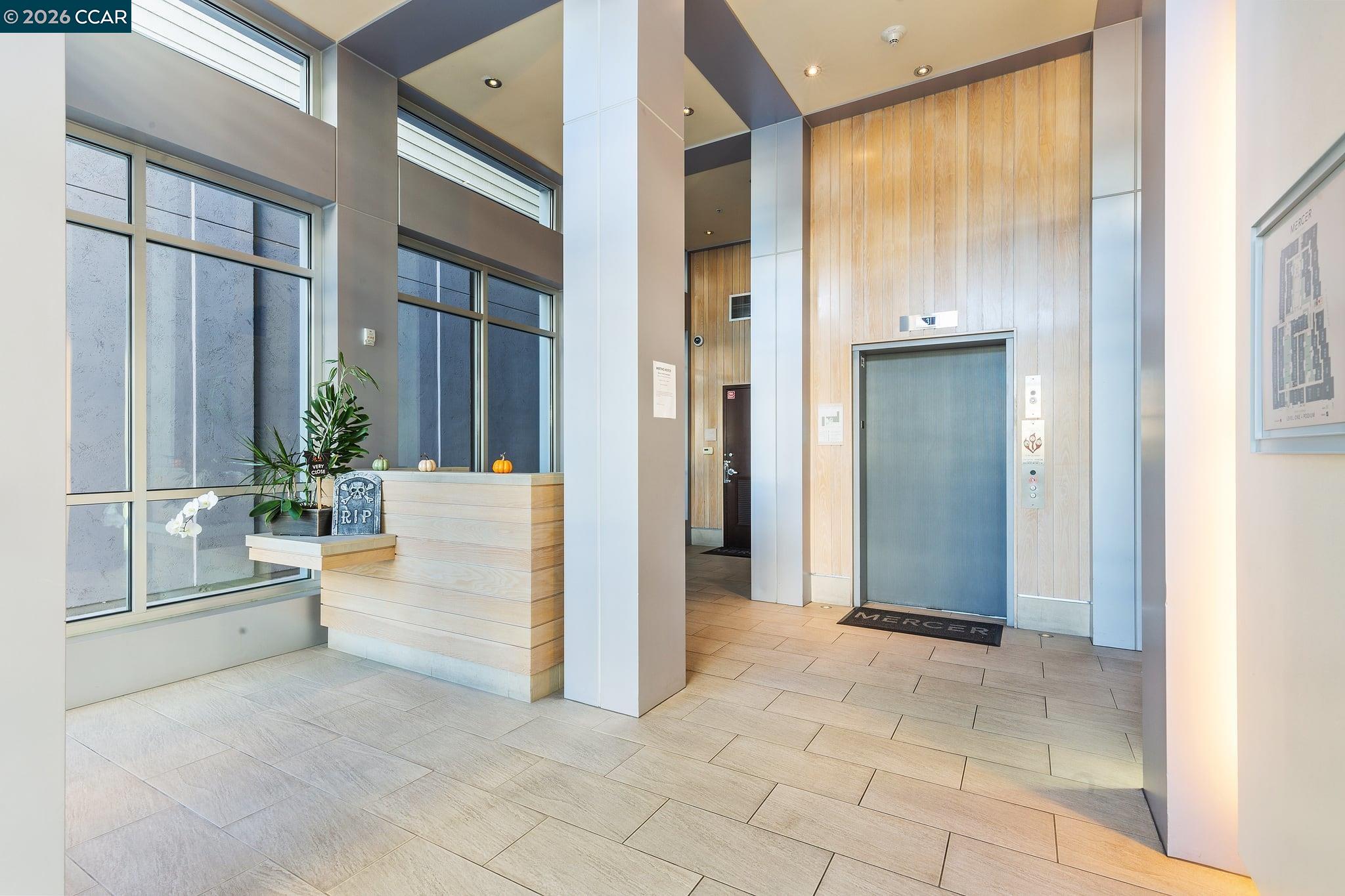 Condominium lobby with a wood-paneled elevator, tall windows, and a plant-decorated reception counter.