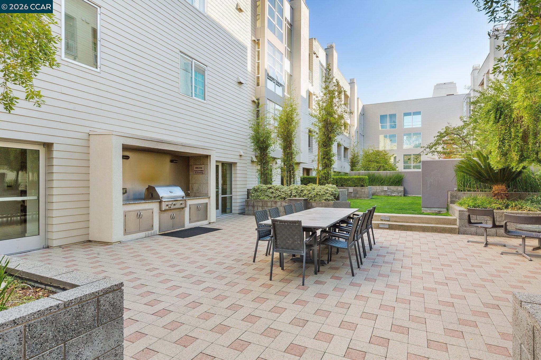 Shared residential courtyard with outdoor dining table, built-in barbecue, and landscaped planters.