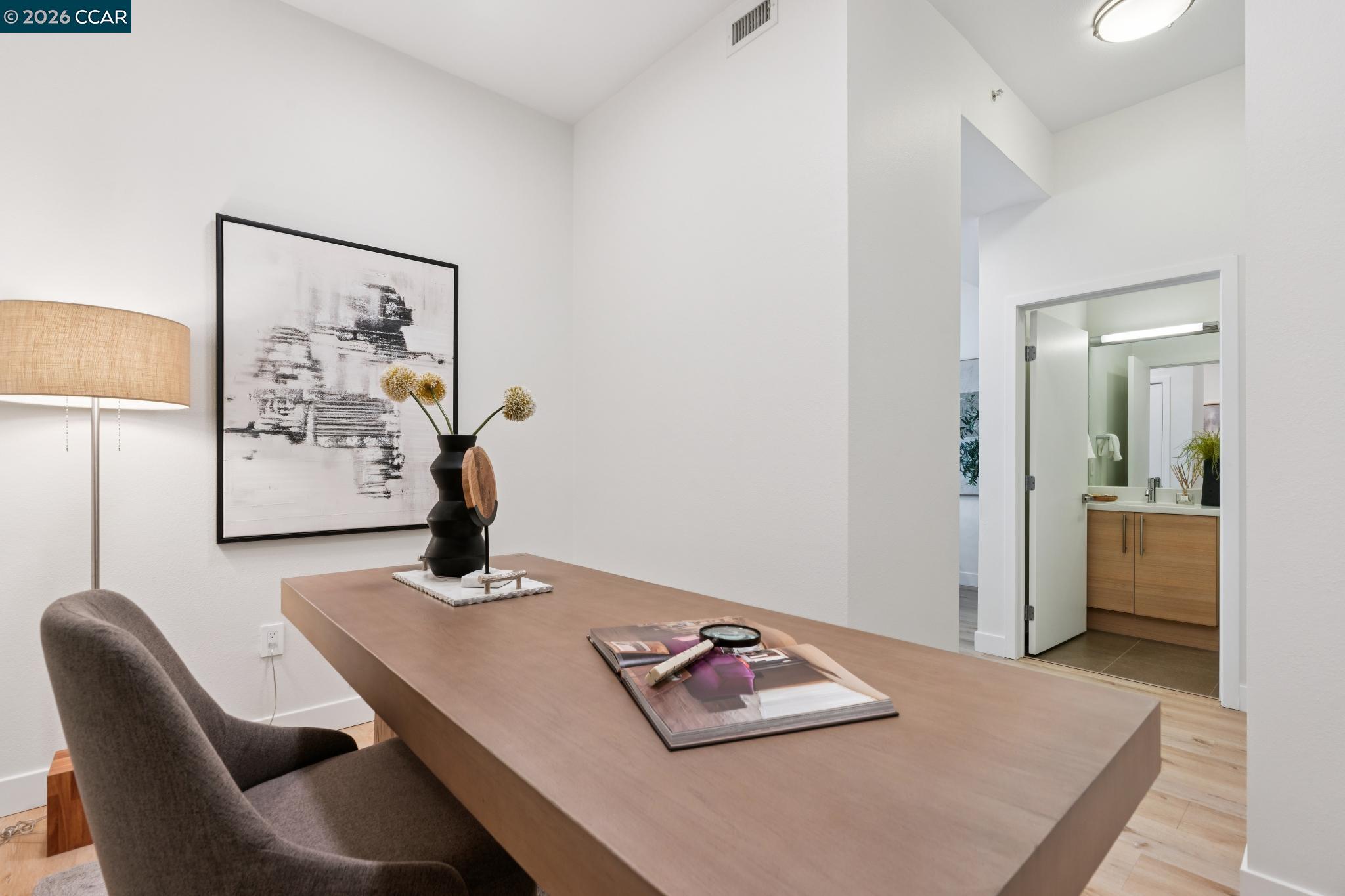 Staged home office with a broad wooden desk and chair; an open doorway reveals a bathroom vanity.
