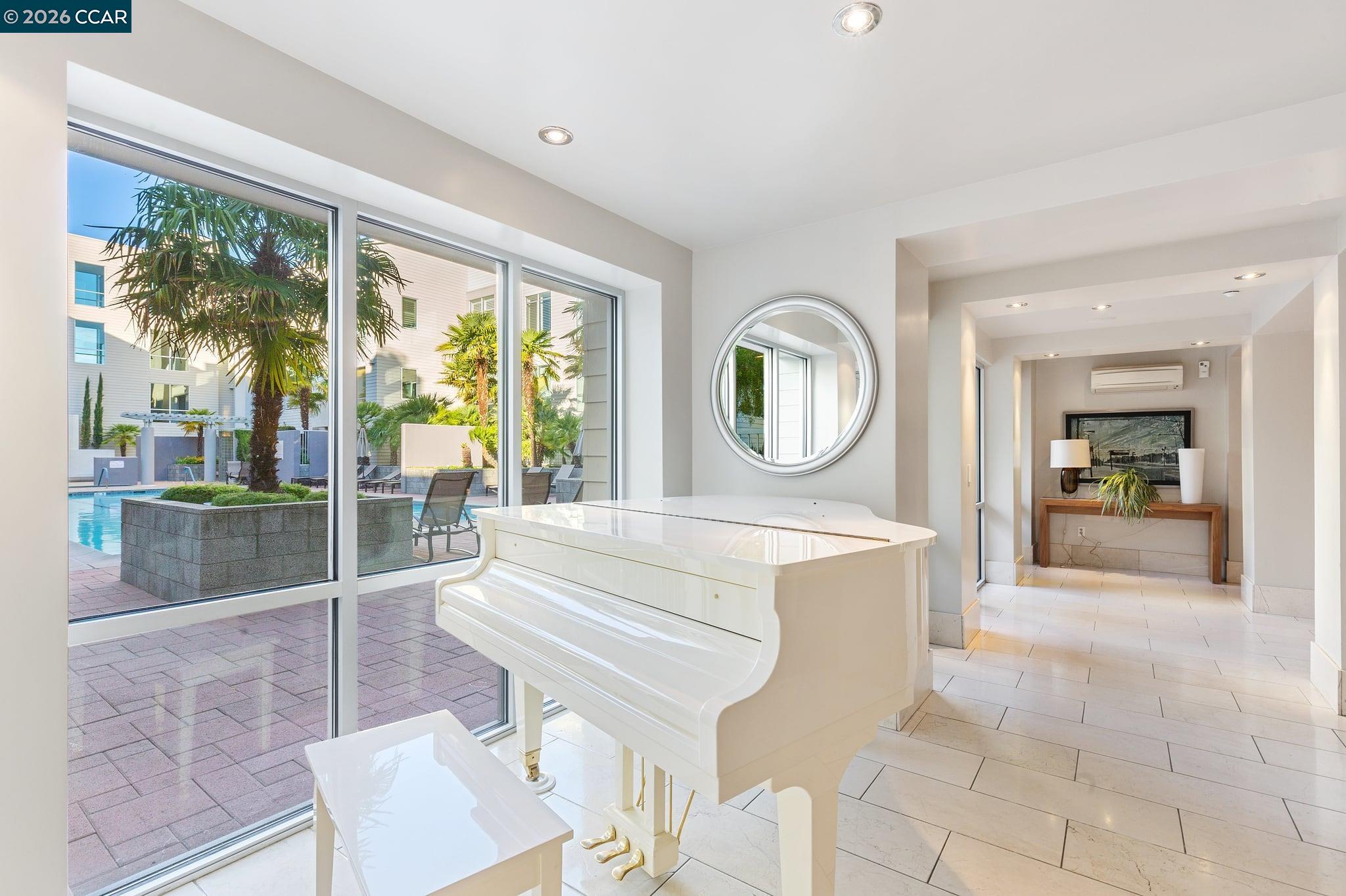 Bright condo living area with a white grand piano overlooking a palm-lined courtyard pool.