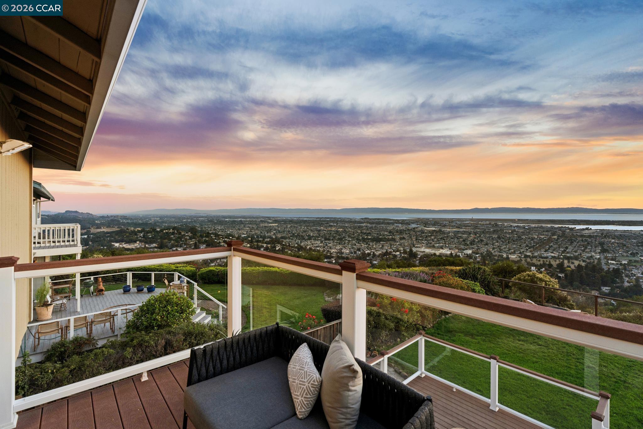 Furnished deck with glass railings overlooking the city and water at sunset.