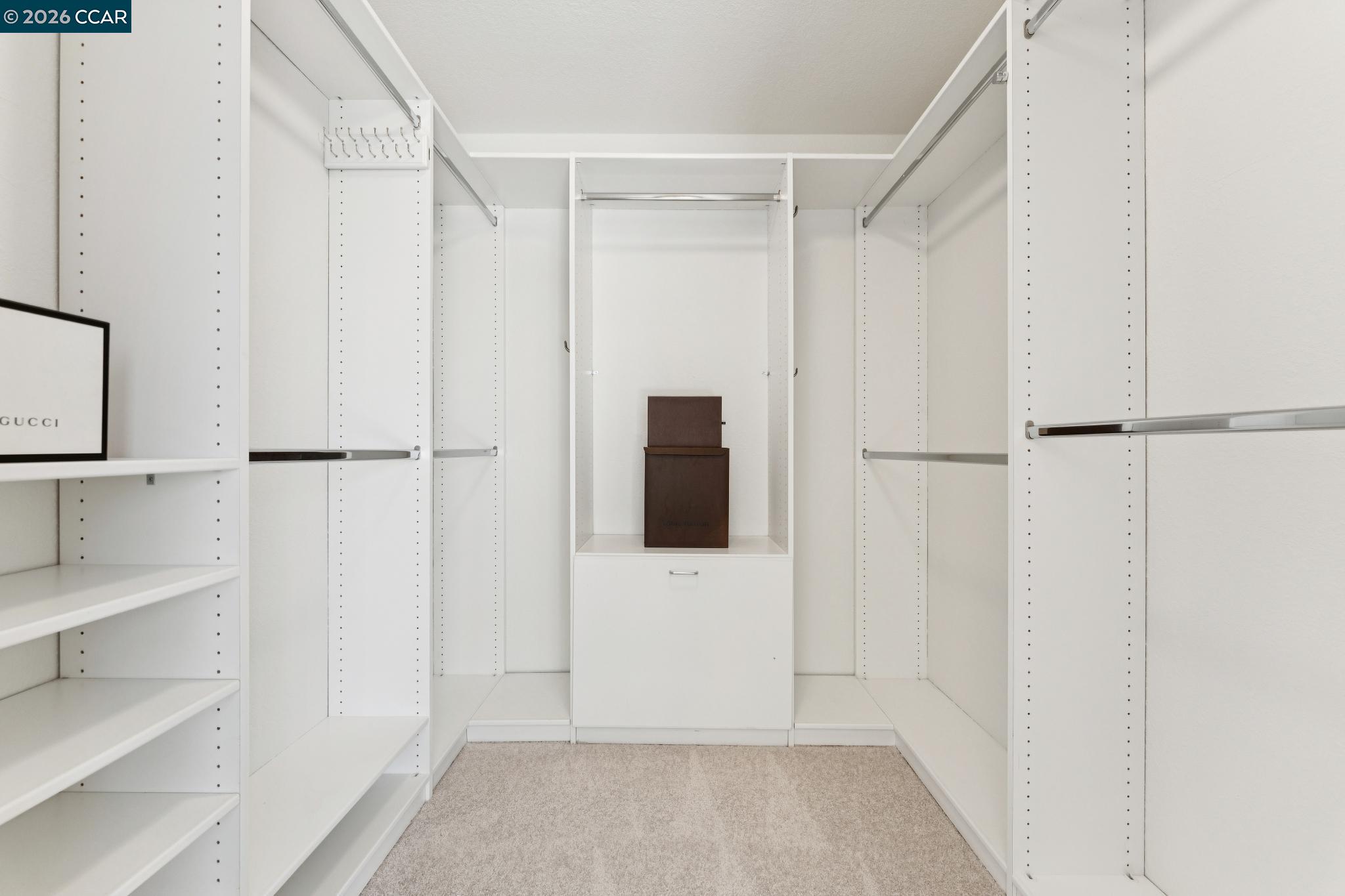 Walk-in closet with white built-in shelves, hanging rods, a central drawer cabinet, and beige carpet.