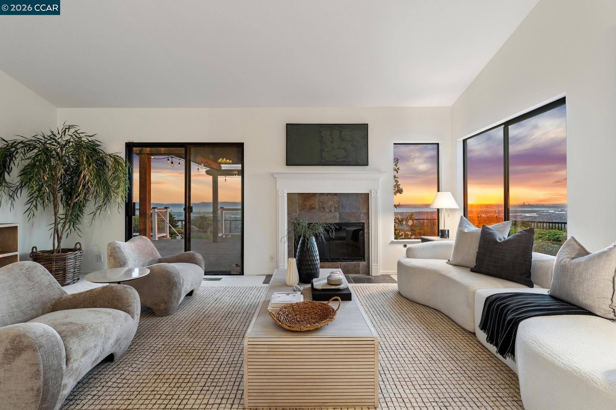 Living room with a fireplace, white sofas, and large windows overlooking water at sunset.