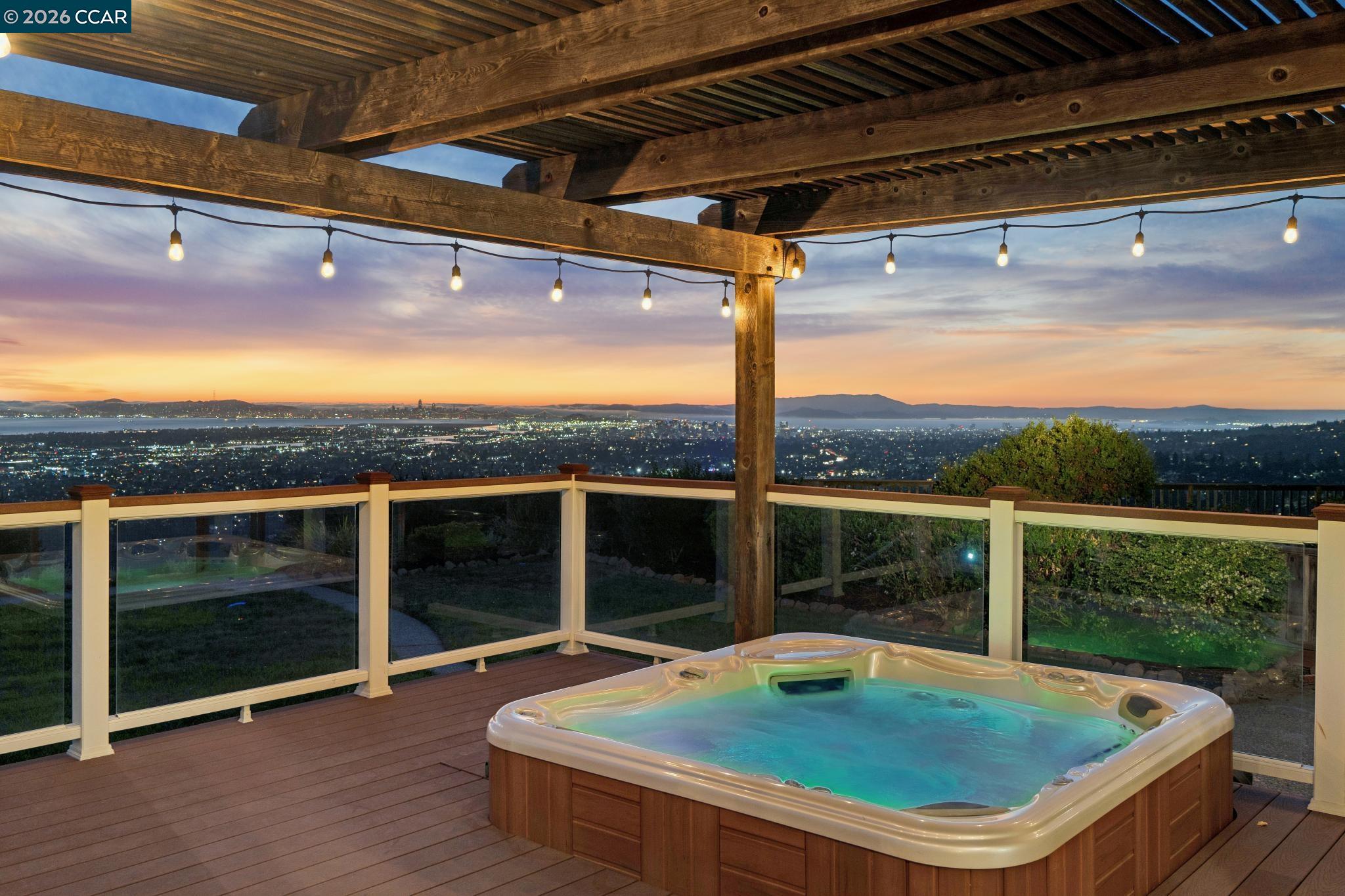 Deck hot tub beneath a timber pergola, overlooking city lights and distant hills at sunset.