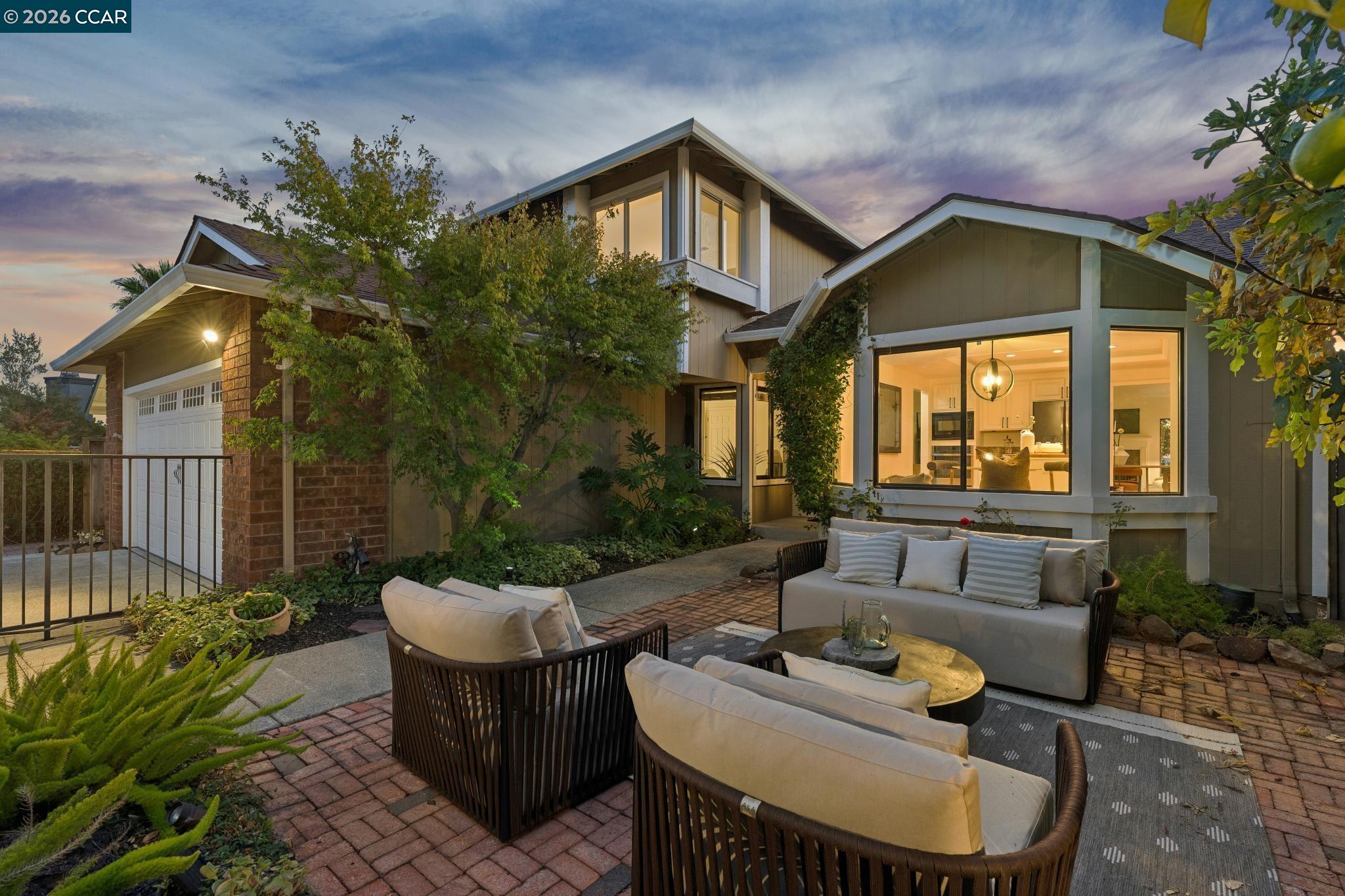 Two-story home at dusk with a brick patio, outdoor seating, and warmly lit windows.