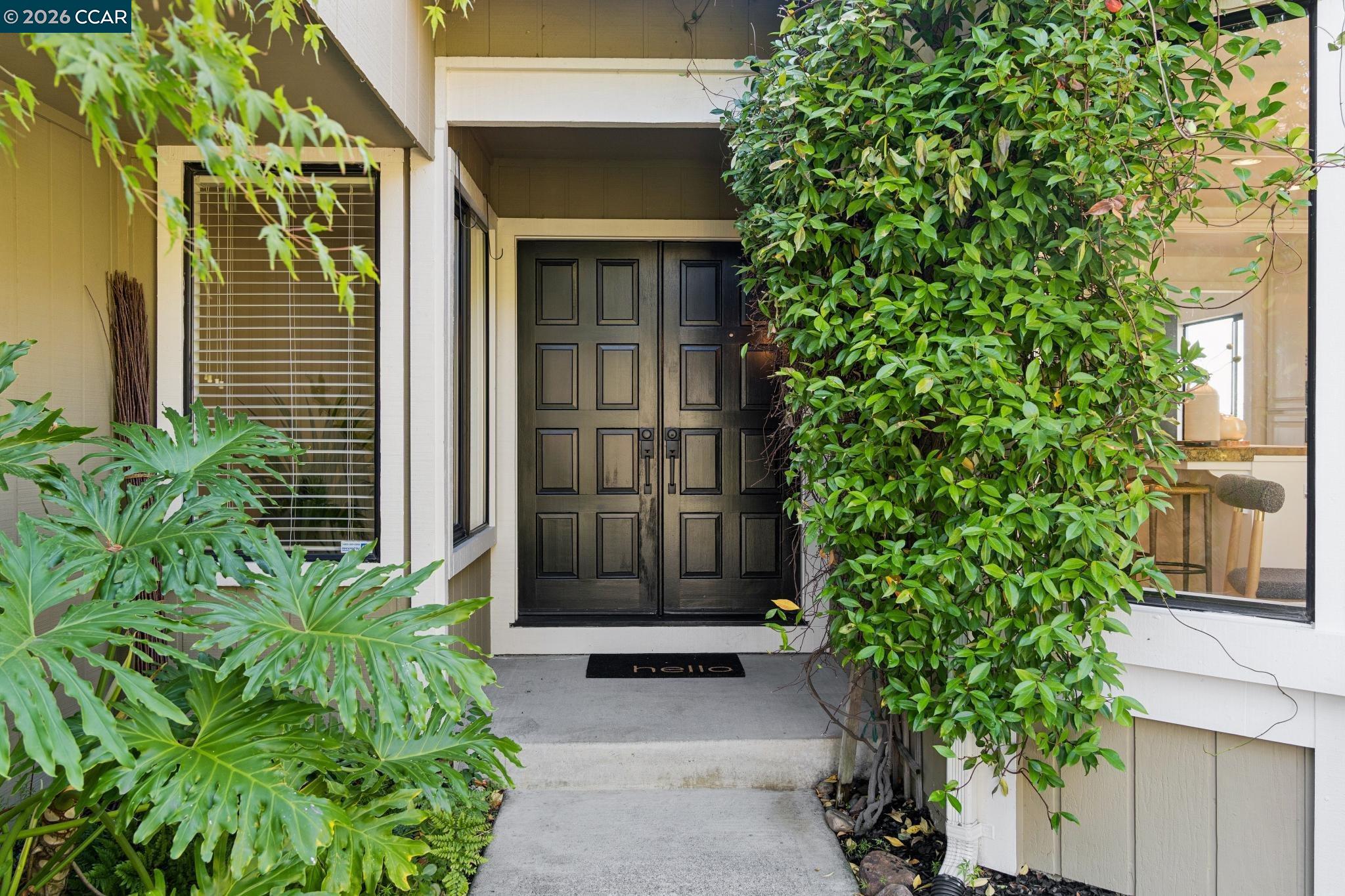 Front entrance with black double doors framed by lush foliage and a concrete walkway.
