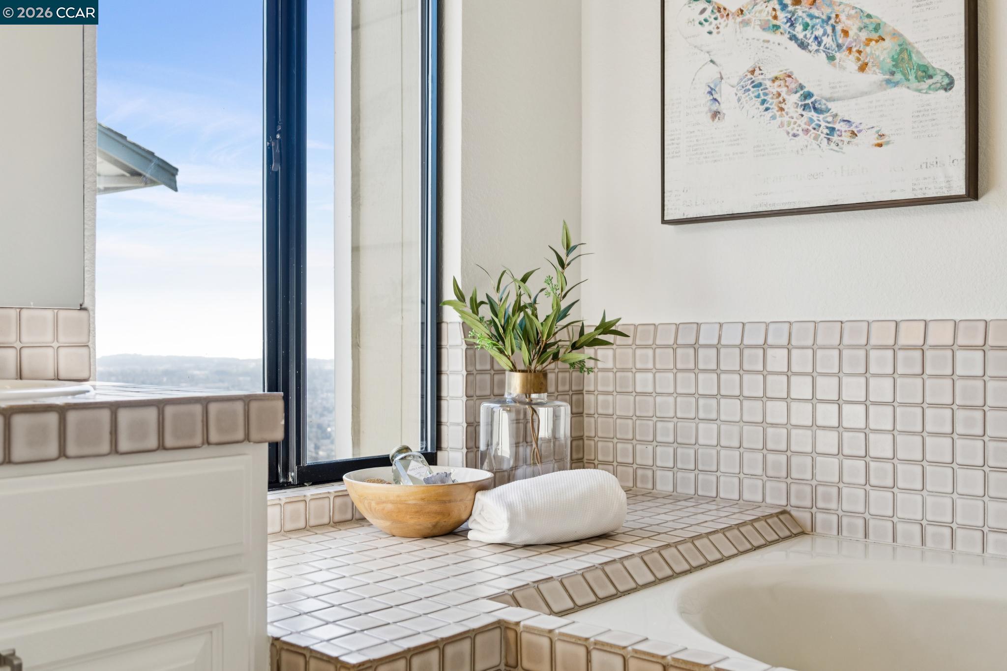 Bathroom with a tiled tub surround, large window, and greenery beside folded towels.