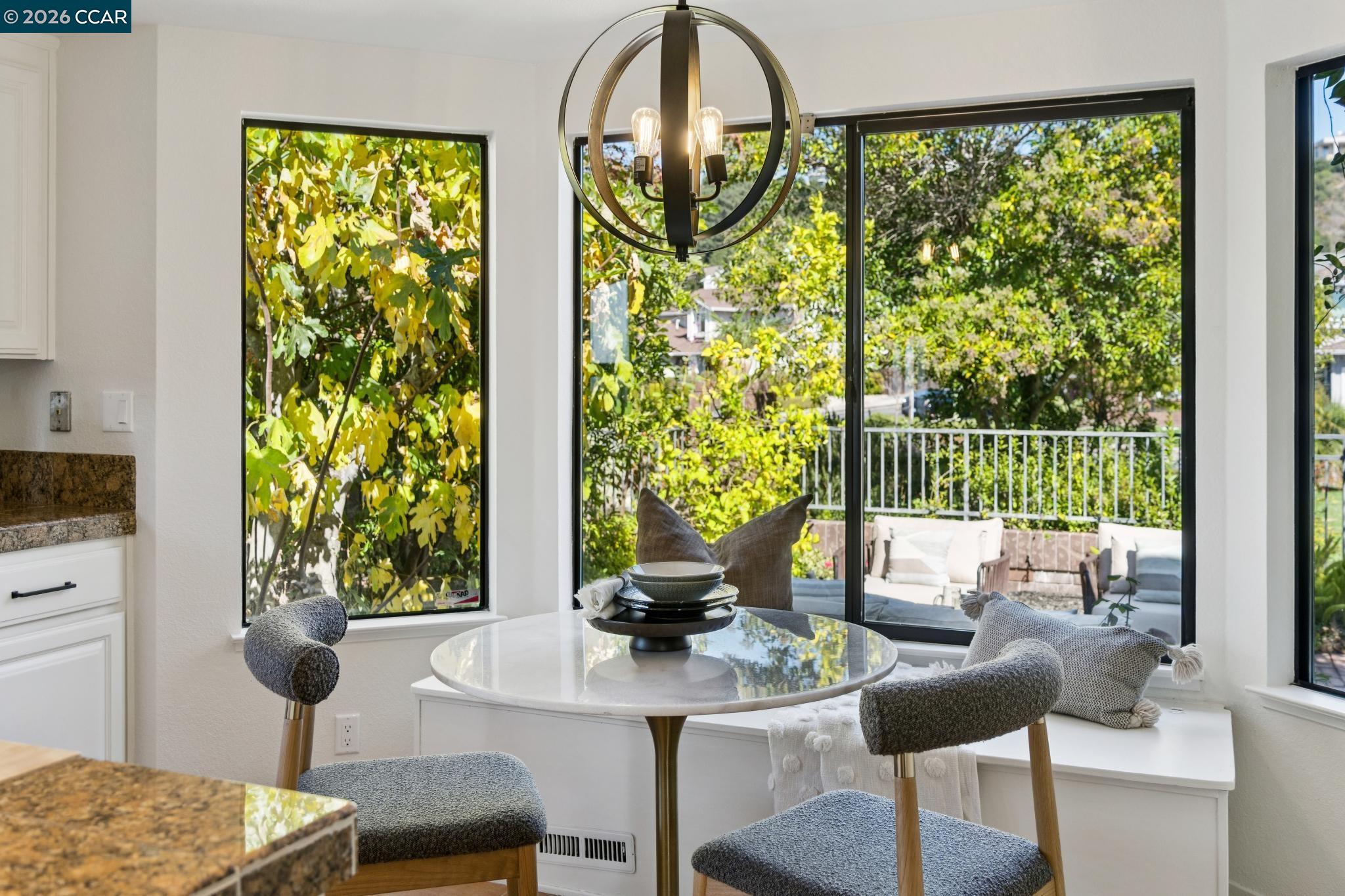 Dining nook with a round table and gray chairs beside large windows overlooking trees and a patio.
