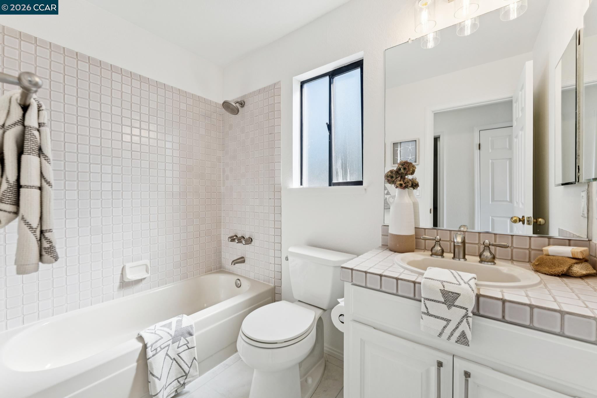 Bathroom with a square-tile tub surround, white vanity, large mirror, and narrow window.