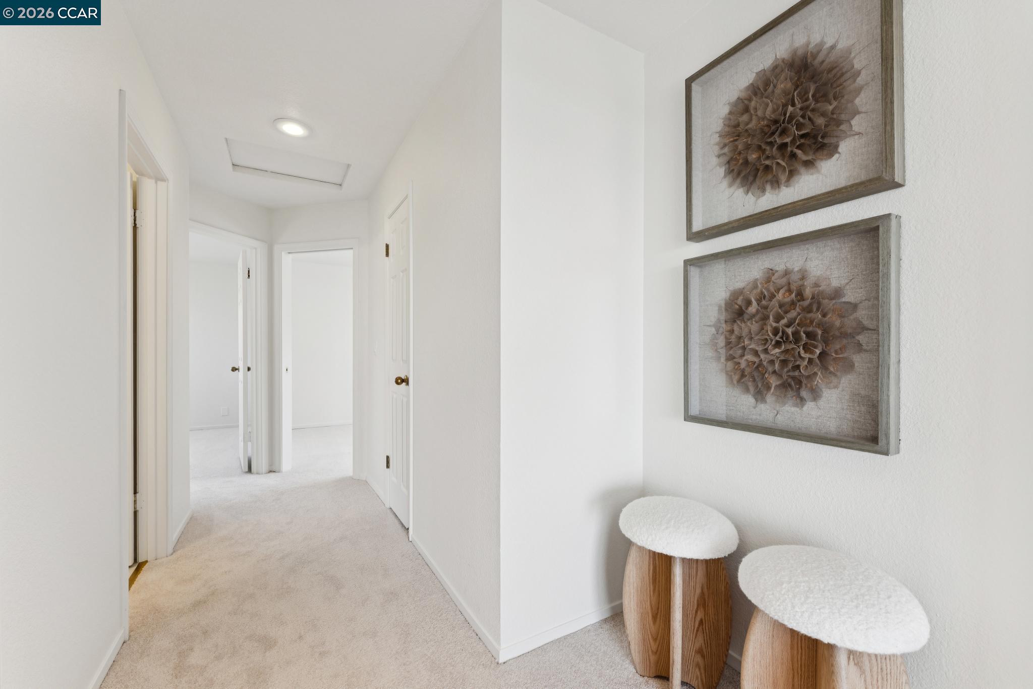 Bright carpeted hallway with open doorways, framed botanical art, and two upholstered stools.
