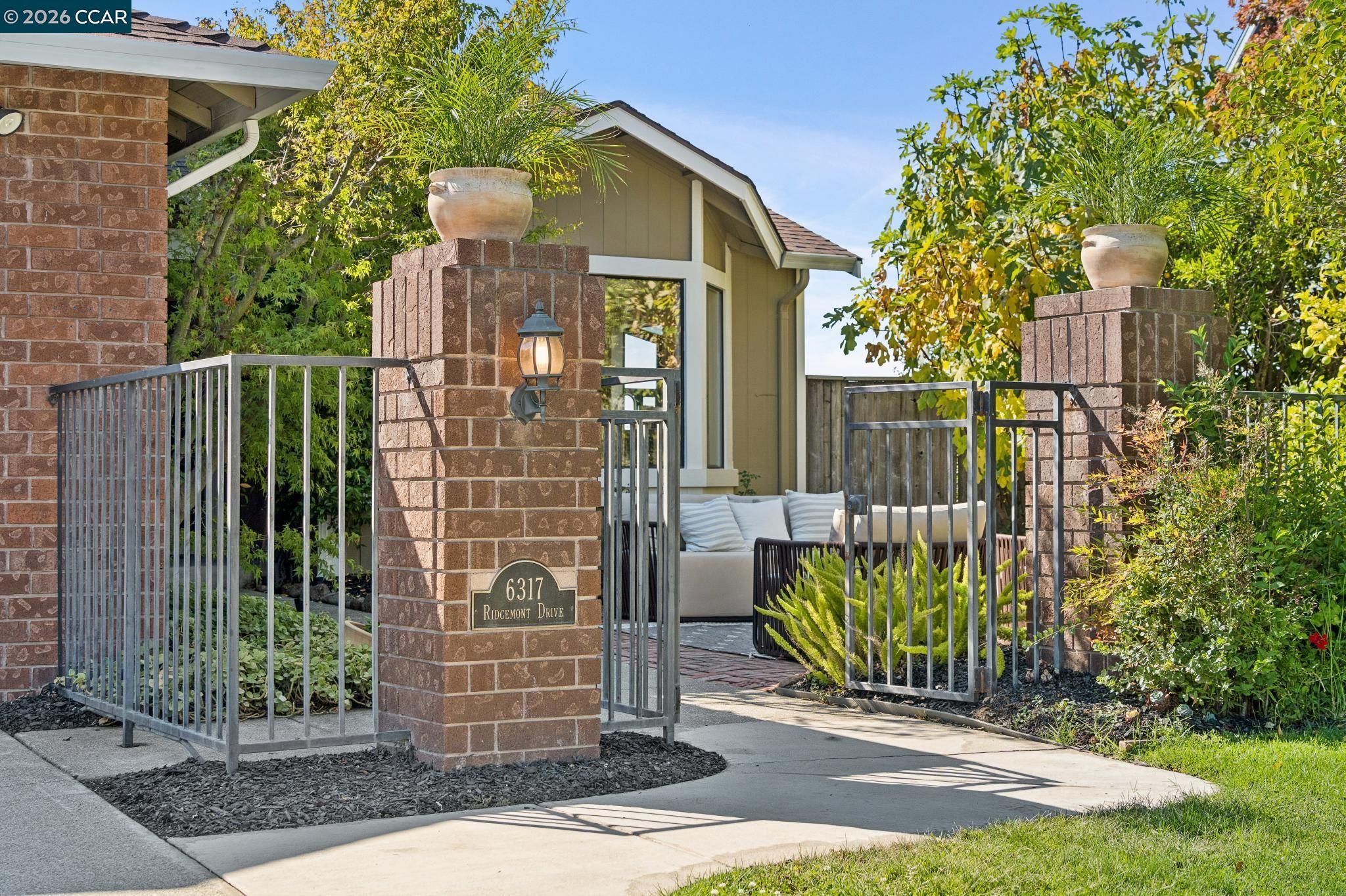 Landscaped home entrance with brick pillars, metal gates, and a seating area beyond.