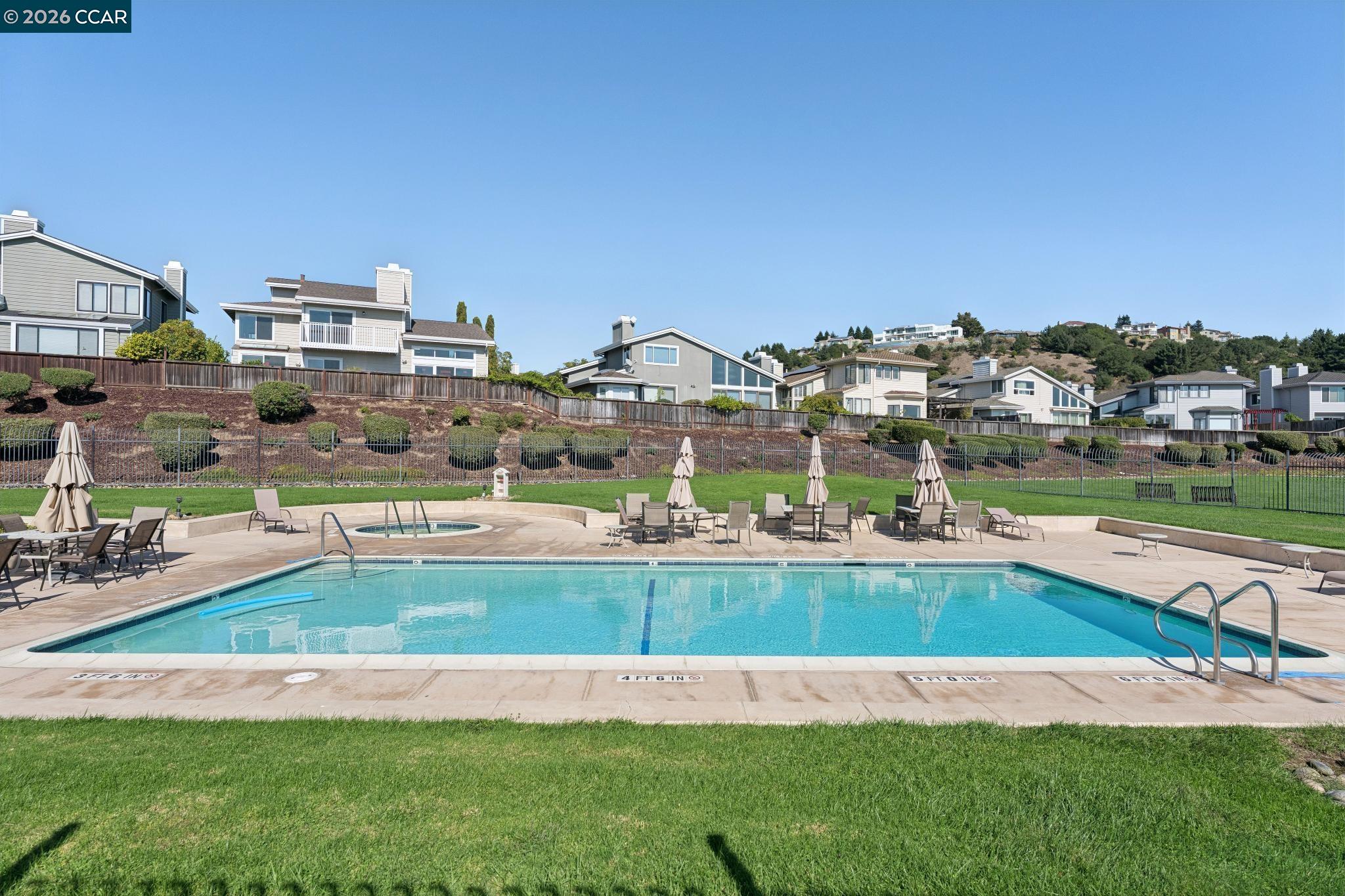 Outdoor swimming pool with a circular spa, deck seating, and hillside homes behind it.