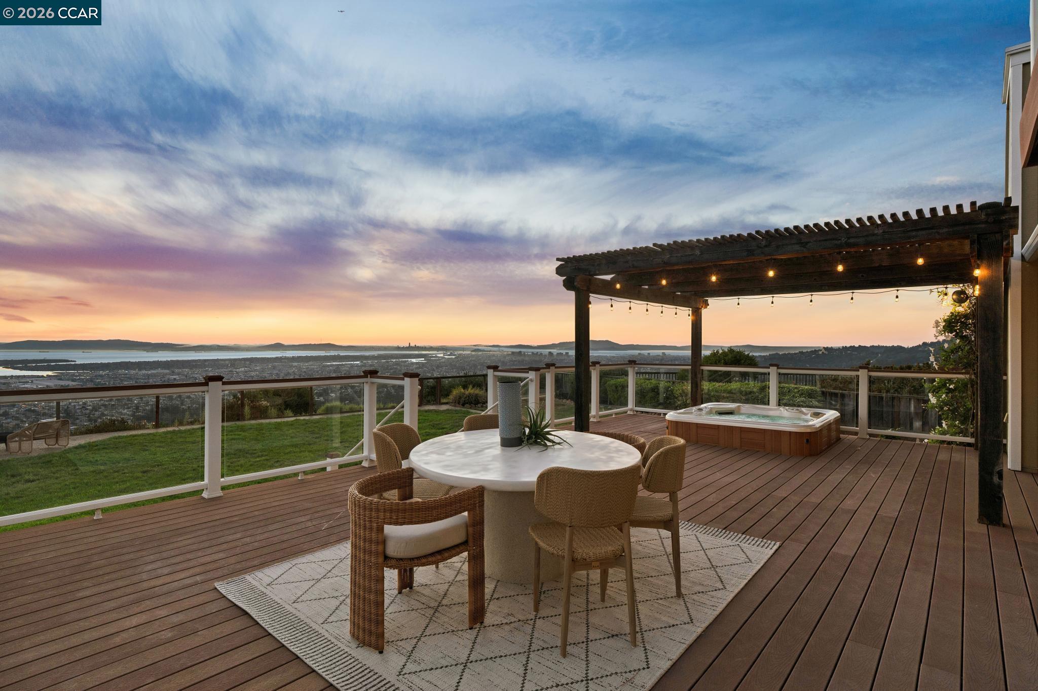 Furnished deck with pergola and hot tub overlooking the water and city at sunset.