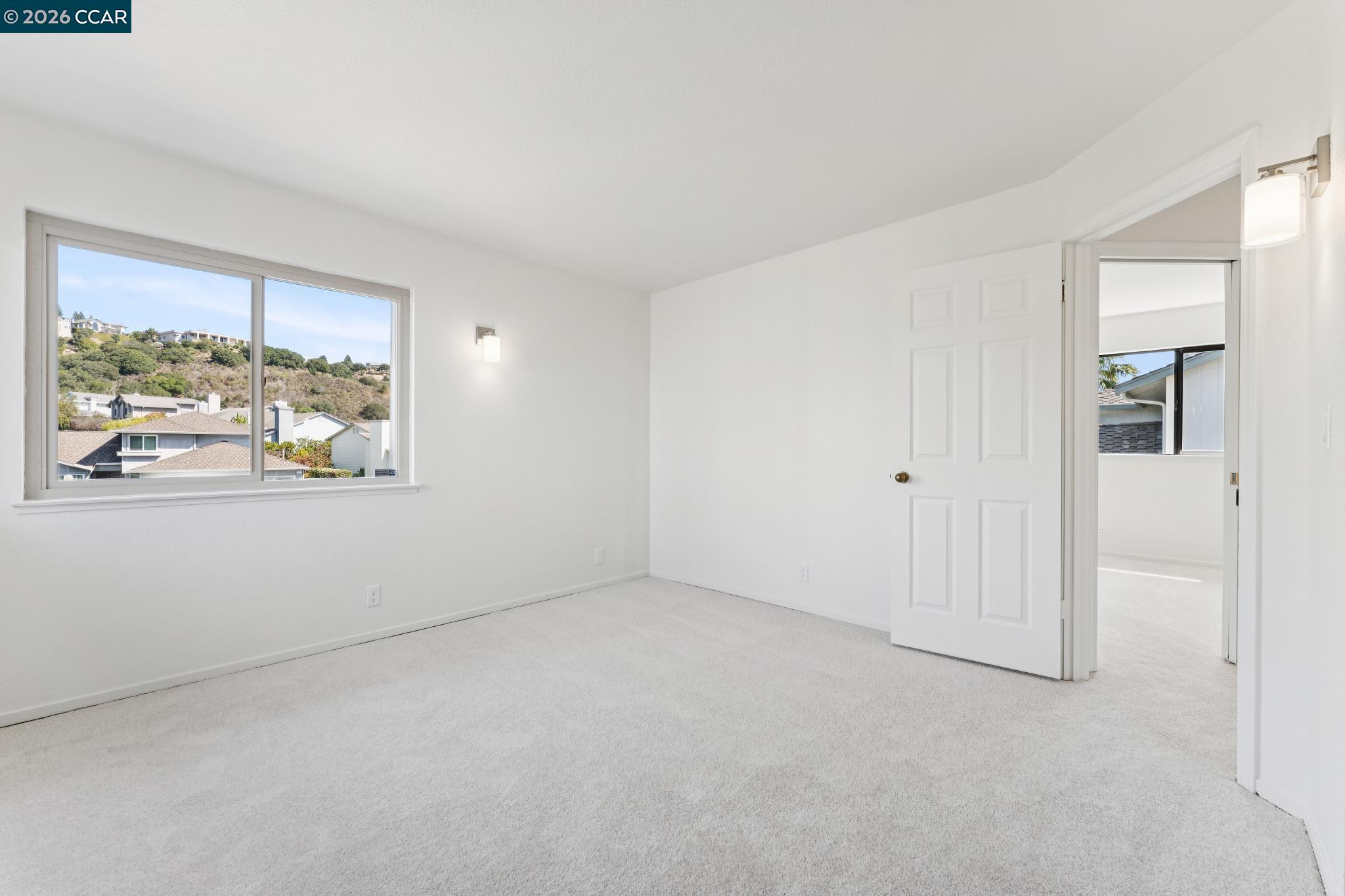 Empty carpeted room with a large window overlooking hillside homes and an open doorway.