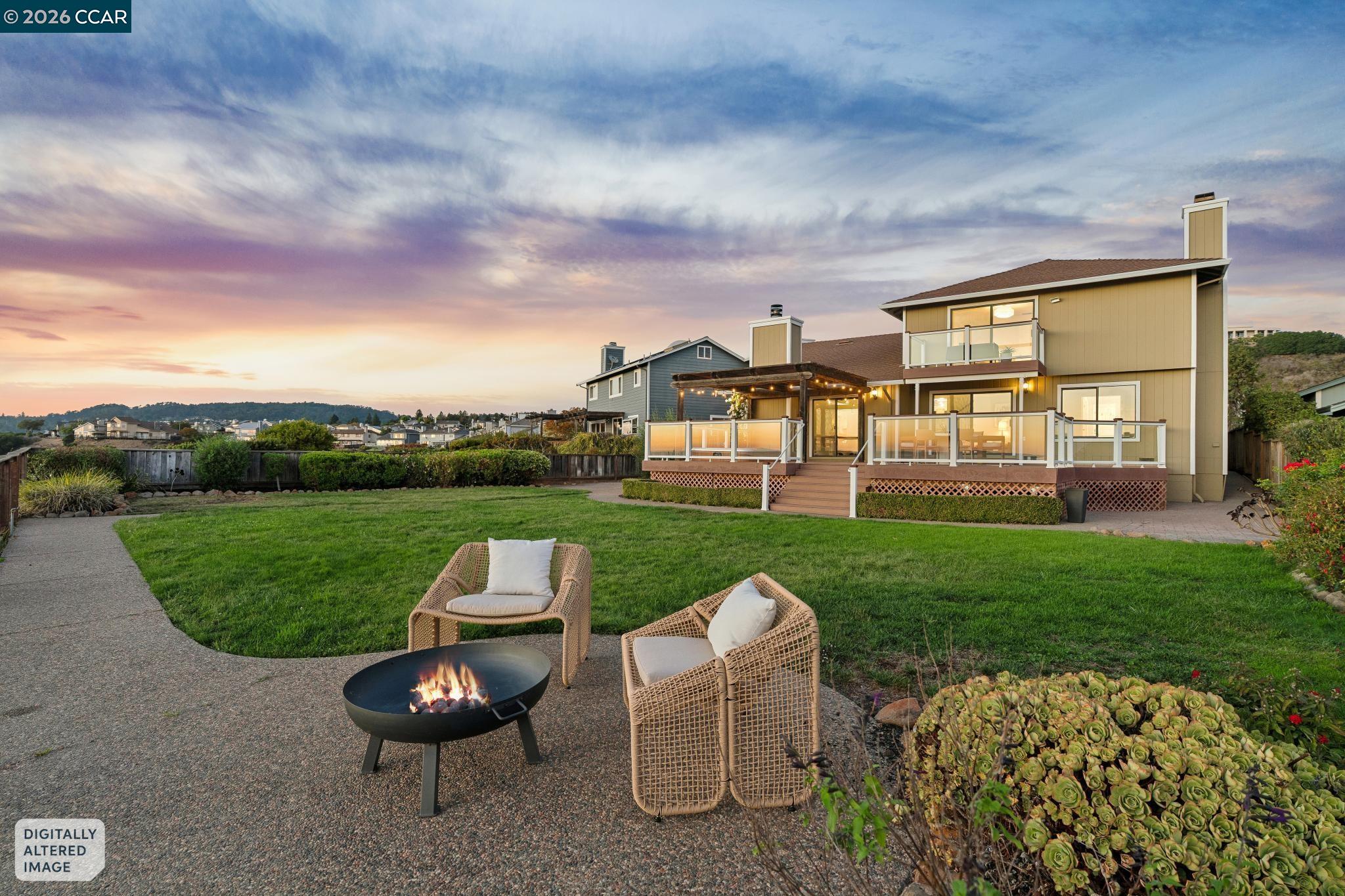 Two-story home behind a lawn, with a raised deck, patio chairs, and a fire pit under a sunset sky.