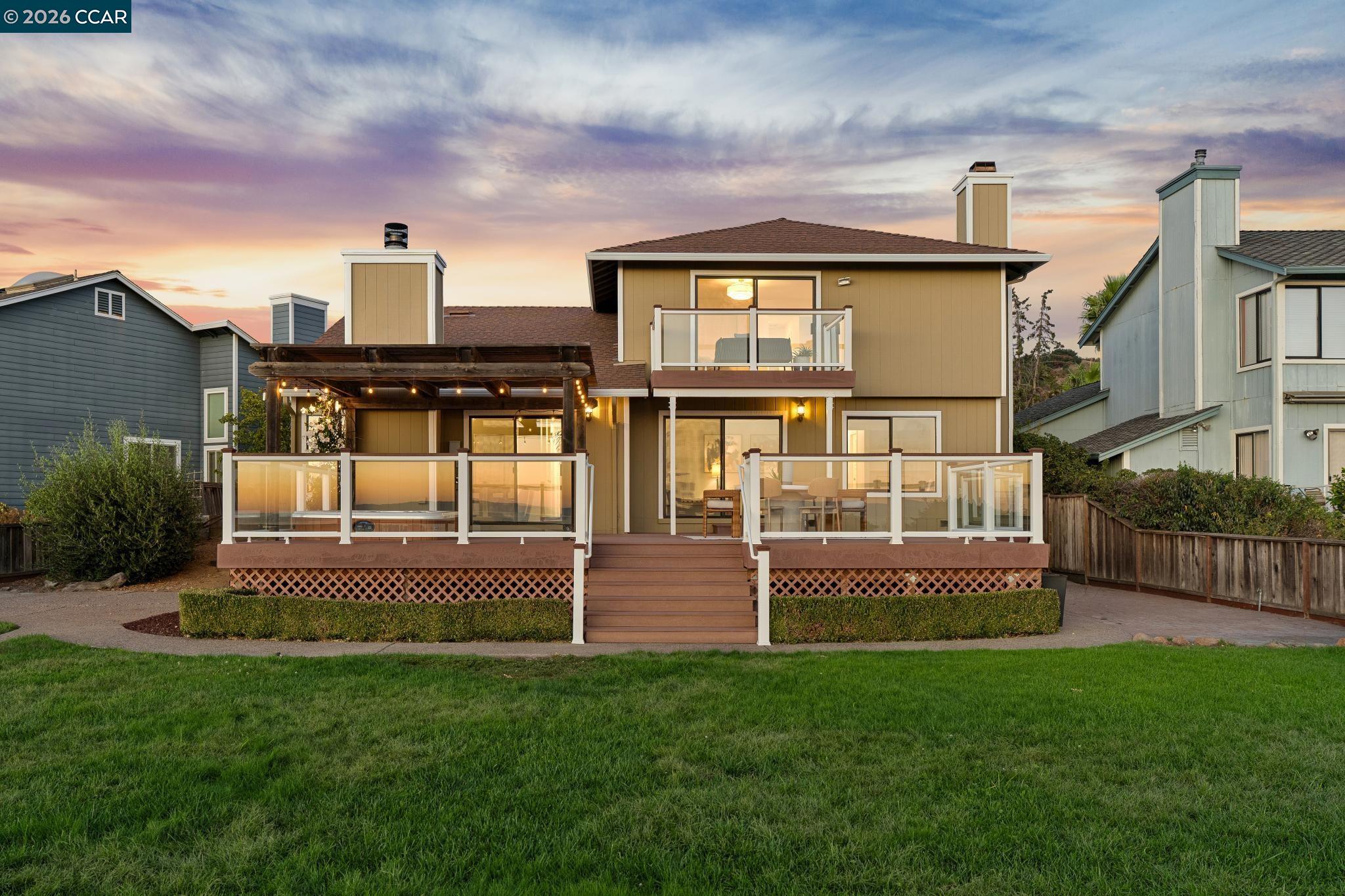 Rear exterior of a two-story home with a broad glass-railed deck and pergola at sunset.