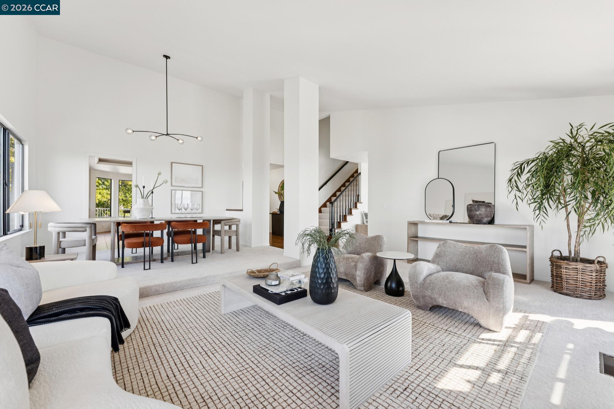 Staged living and dining room with light seating, rust-colored dining chairs, large windows, and a staircase.