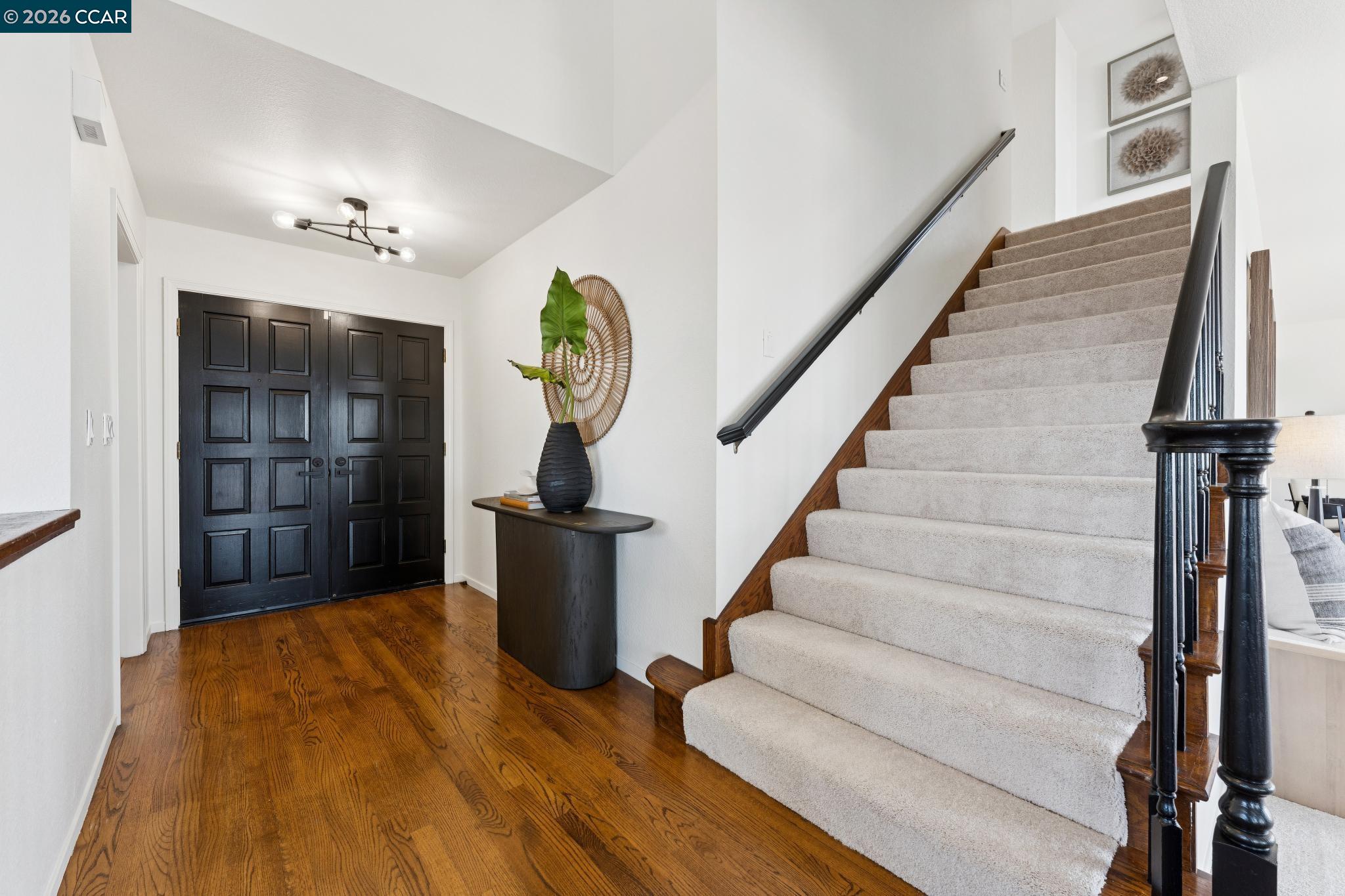 Entry foyer with dark double doors, hardwood floors, and a carpeted staircase with dark railings.